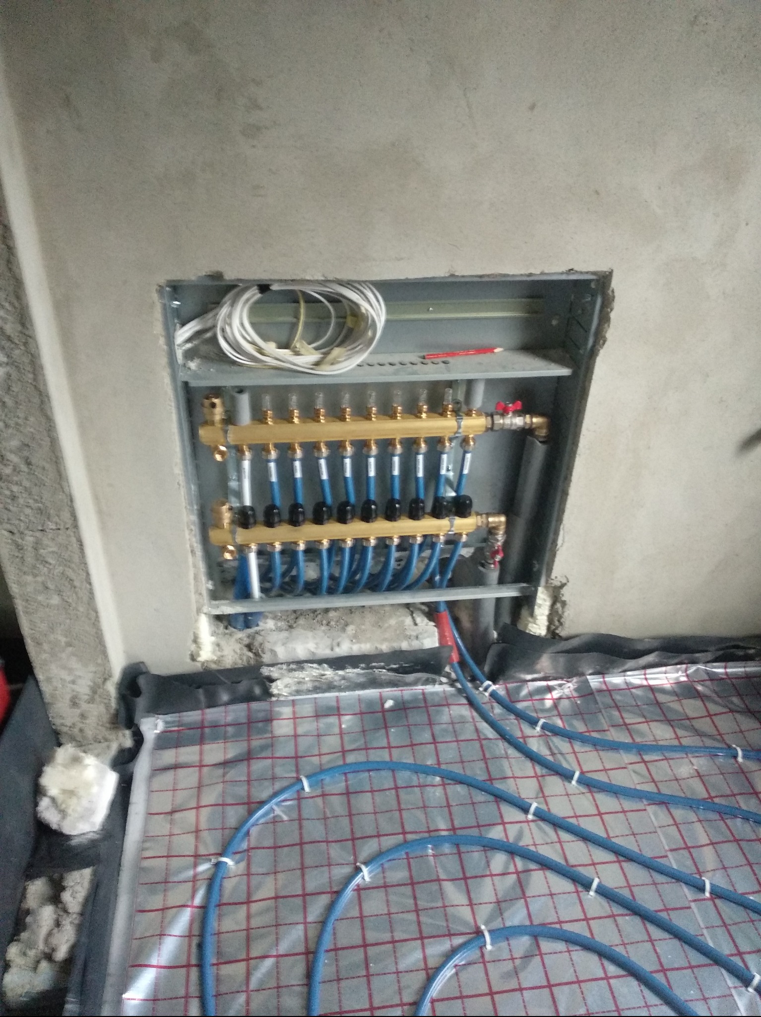 Rozdzielacz ogrzewania podłogowego z niebieskimi rurami na folii z czerwoną siatką, zamontowany w szarej wnęce w ścianie. Widoczne przewody i ołówek.