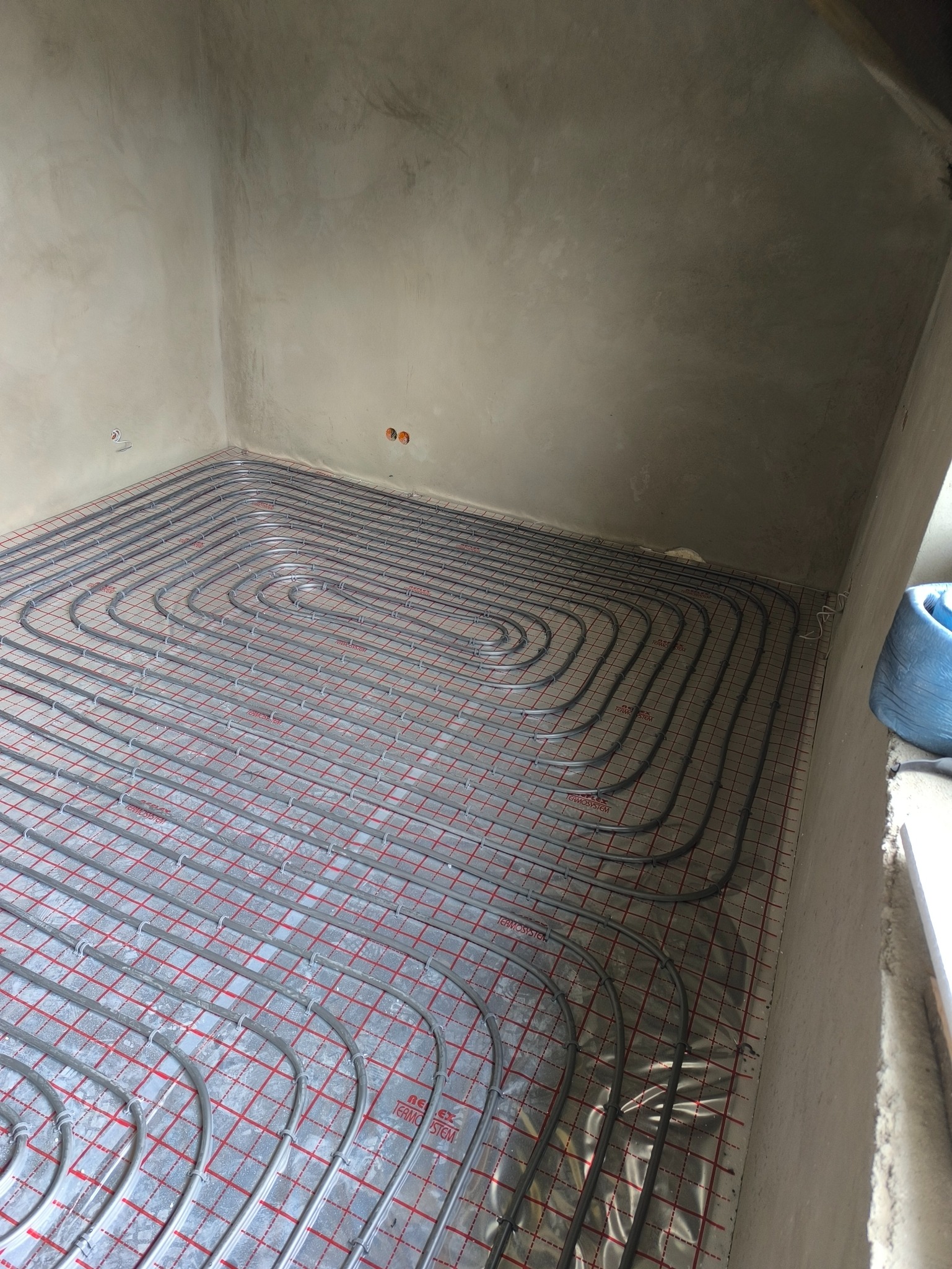 Szare rury ogrzewania podłogowego ułożone na folii z czerwoną siatką w surowym pomieszczeniu. Widoczne dwa otwory w ścianie. Ujęcie z góry.