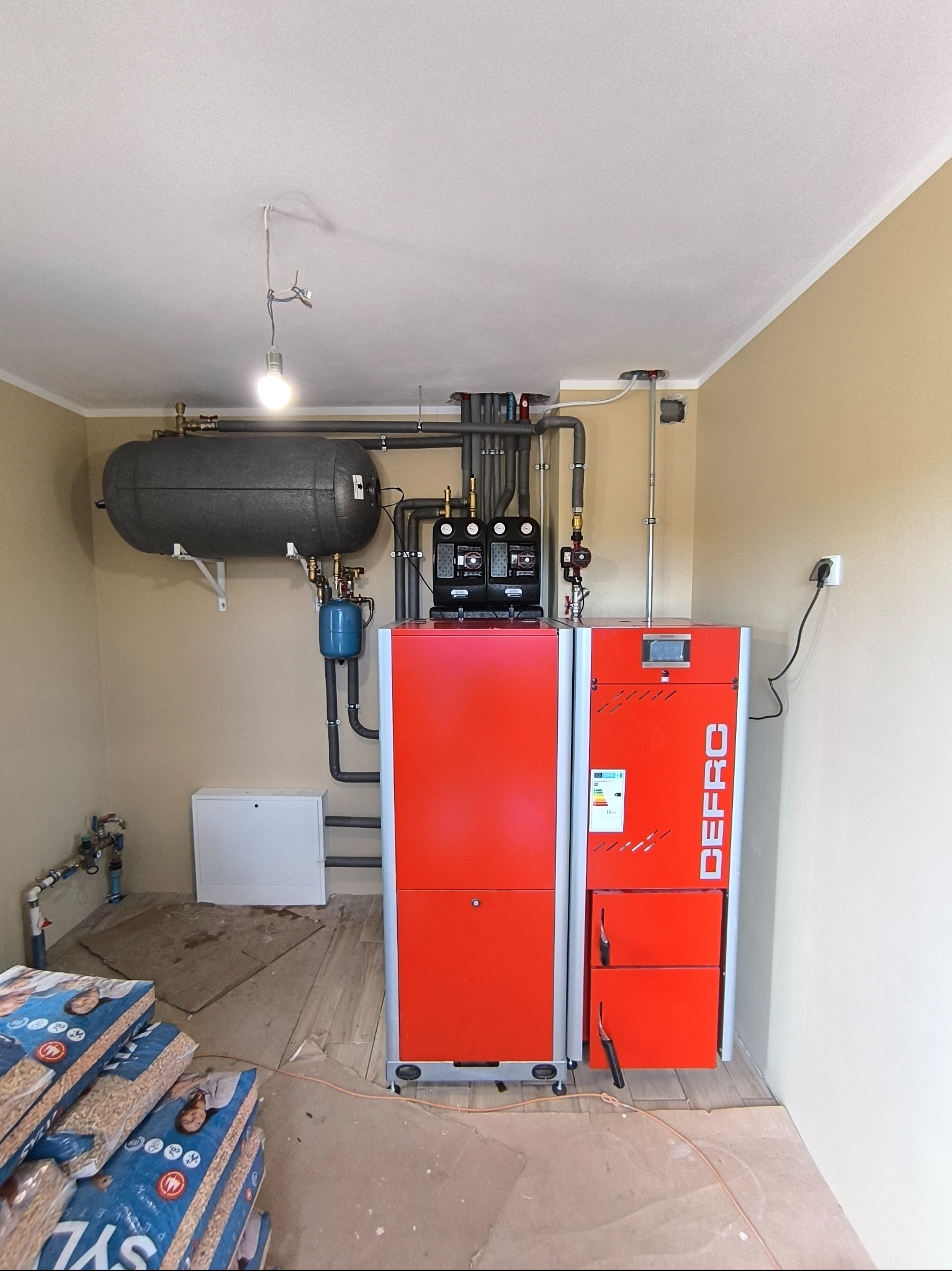 Kotłownia z czerwonym piecem na pellet Defro, zbiornikiem akumulacyjnym i instalacją. Worki z pelletem na podłodze. Widok na kompletną instalację grzewczą w pomieszczeniu.