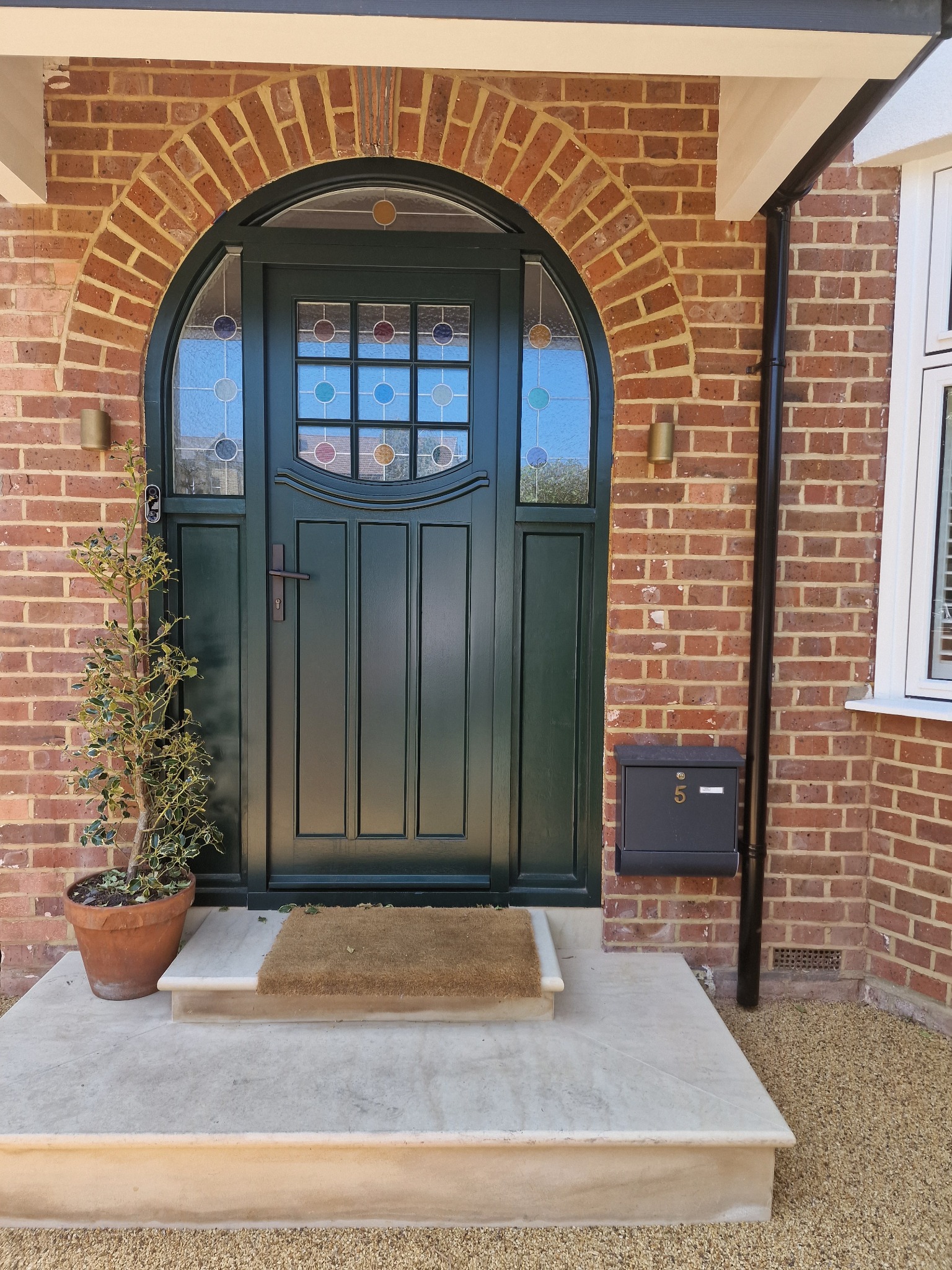 Zielone drzwi wejściowe z łukowym przeszkleniem w ceglanej fasadzie domu. Donica z rośliną po lewej, skrzynka na listy po prawej. Wyjątkowy detal architektoniczny.