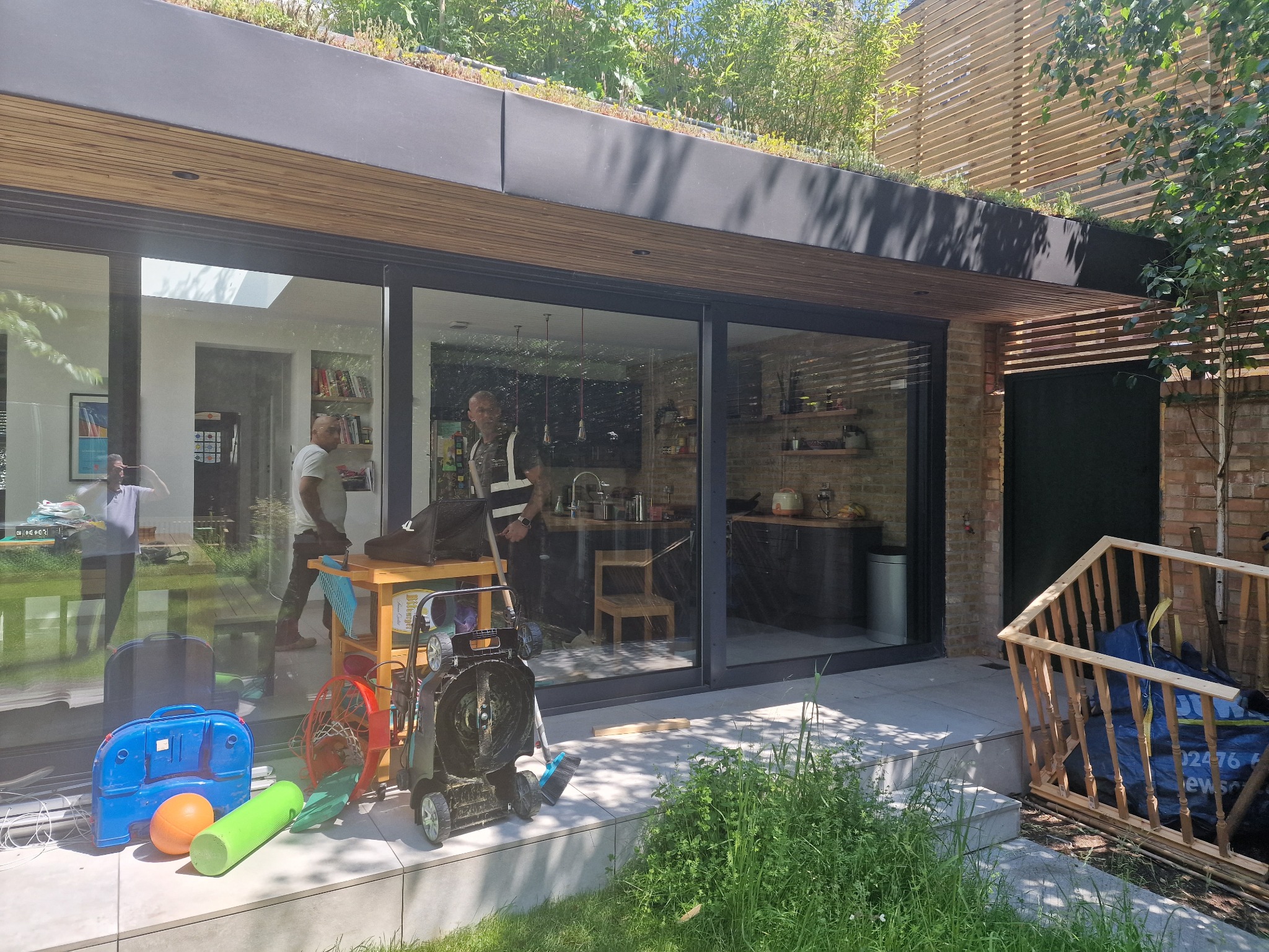 Montaż przesuwnych drzwi tarasowych z czarnymi ramami w nowoczesnym domu z zielonym dachem. Widoczne narzędzia i ekipa montażowa. Częściowe odbicie wnętrza domu.