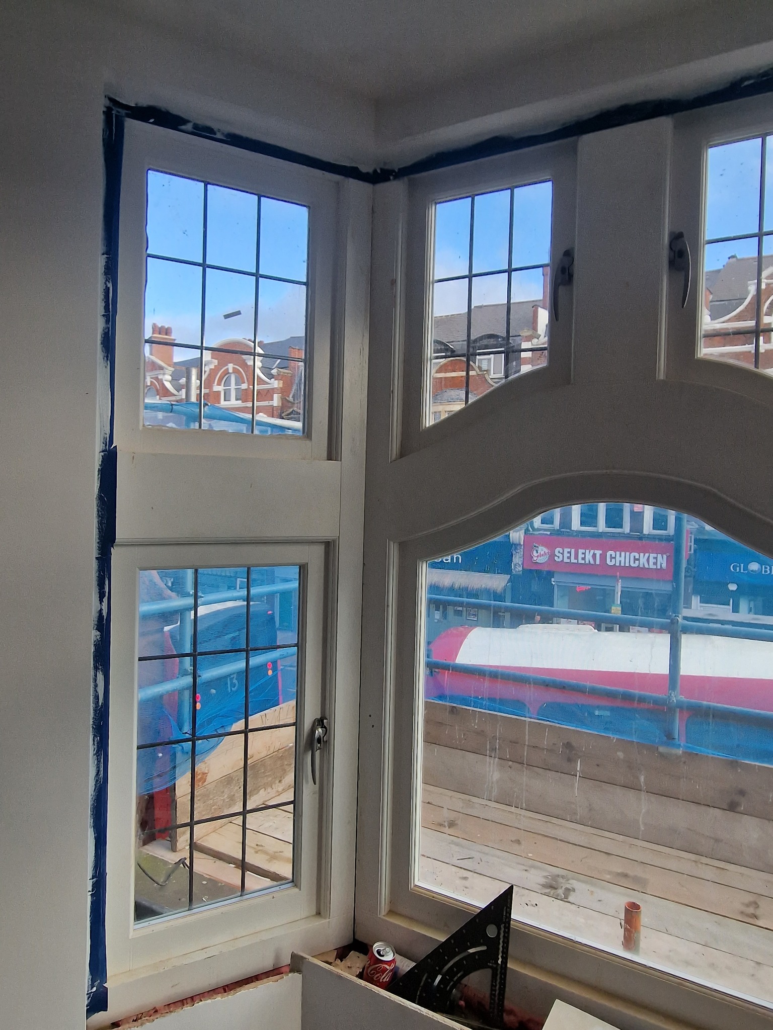 Kątowe okno z białymi ramami, widok na zewnątrz z rusztowaniami i budynkami. W dolnej części okna widoczne narzędzia i puszka napoju.