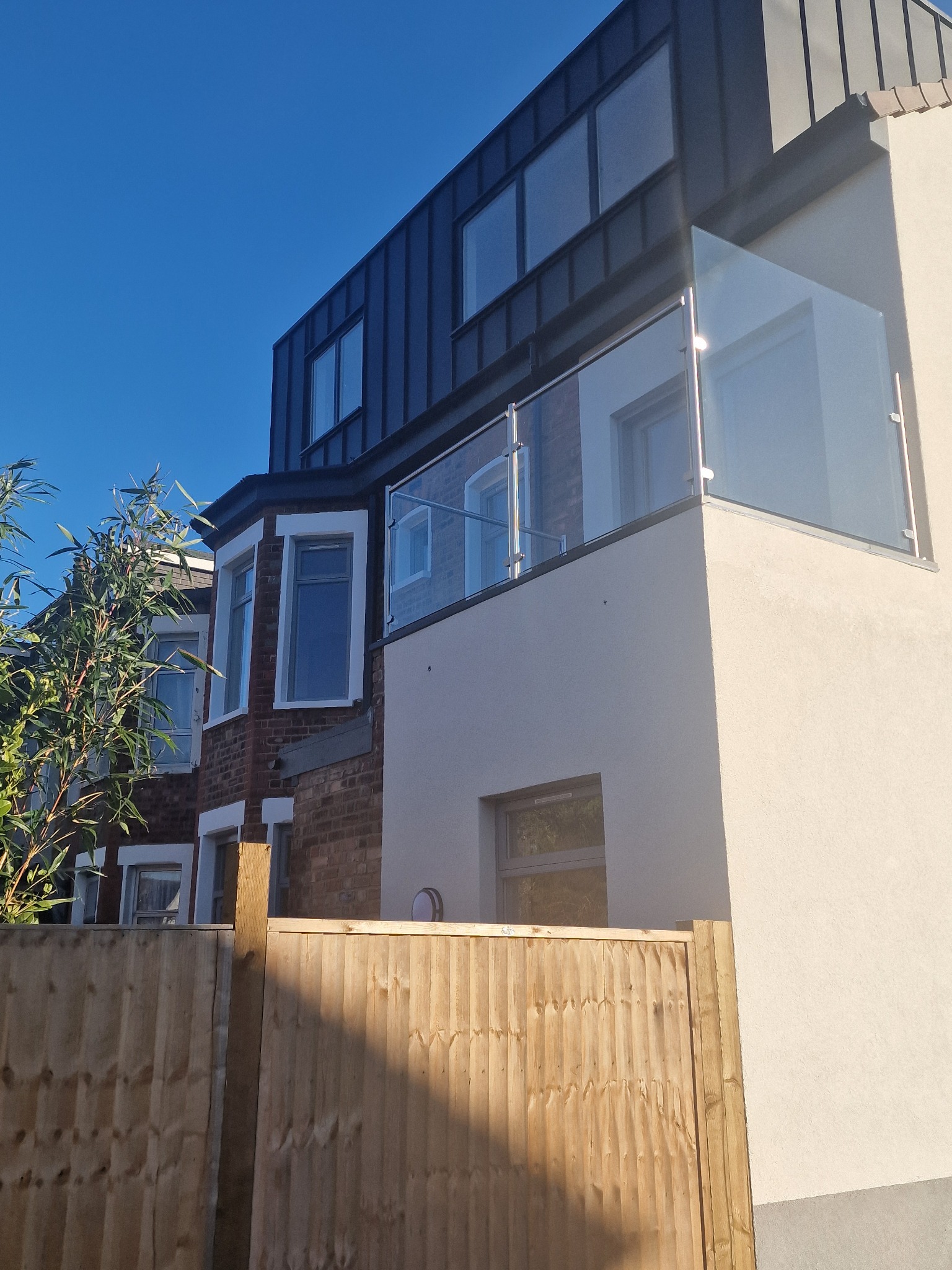 Elewacja domu po remoncie z oknami i balkonem z szklaną balustradą, widok z dołu, na pierwszym planie drewniany płot. Jasne, słoneczne oświetlenie.