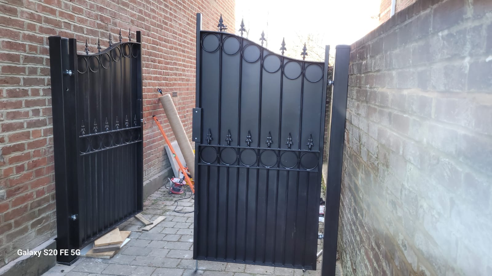 Czarne, metalowe furtki ogrodowe z dekoracyjnymi elementami, zamontowane między ceglanymi murami. Widoczne narzędzia i materiały montażowe.
