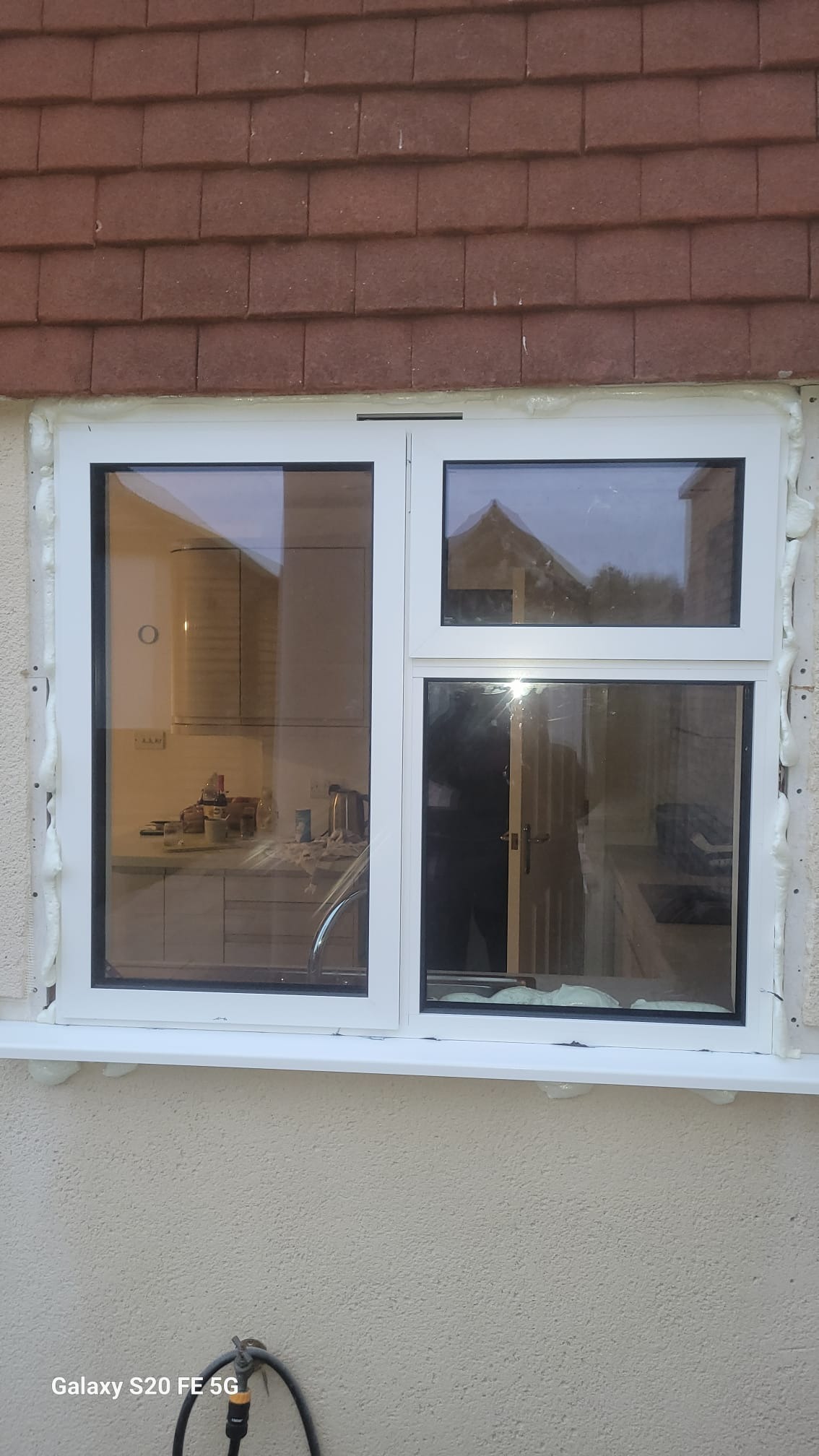 Białe okno PCV z czarnymi uszczelkami, świeżo zamontowane w beżowej ścianie, z widoczną pianką montażową. W tle widoczna kuchnia i odbicie domu w szybie.