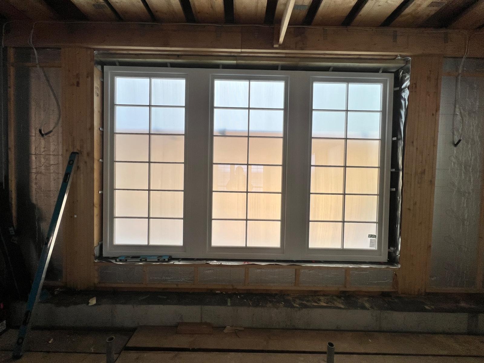 Trzyczęściowe okno z białymi ramami, zamontowane w surowym otworze ściany z drewnianą konstrukcją. Widoczny poziomica i izolacja. W tle rozproszone światło.