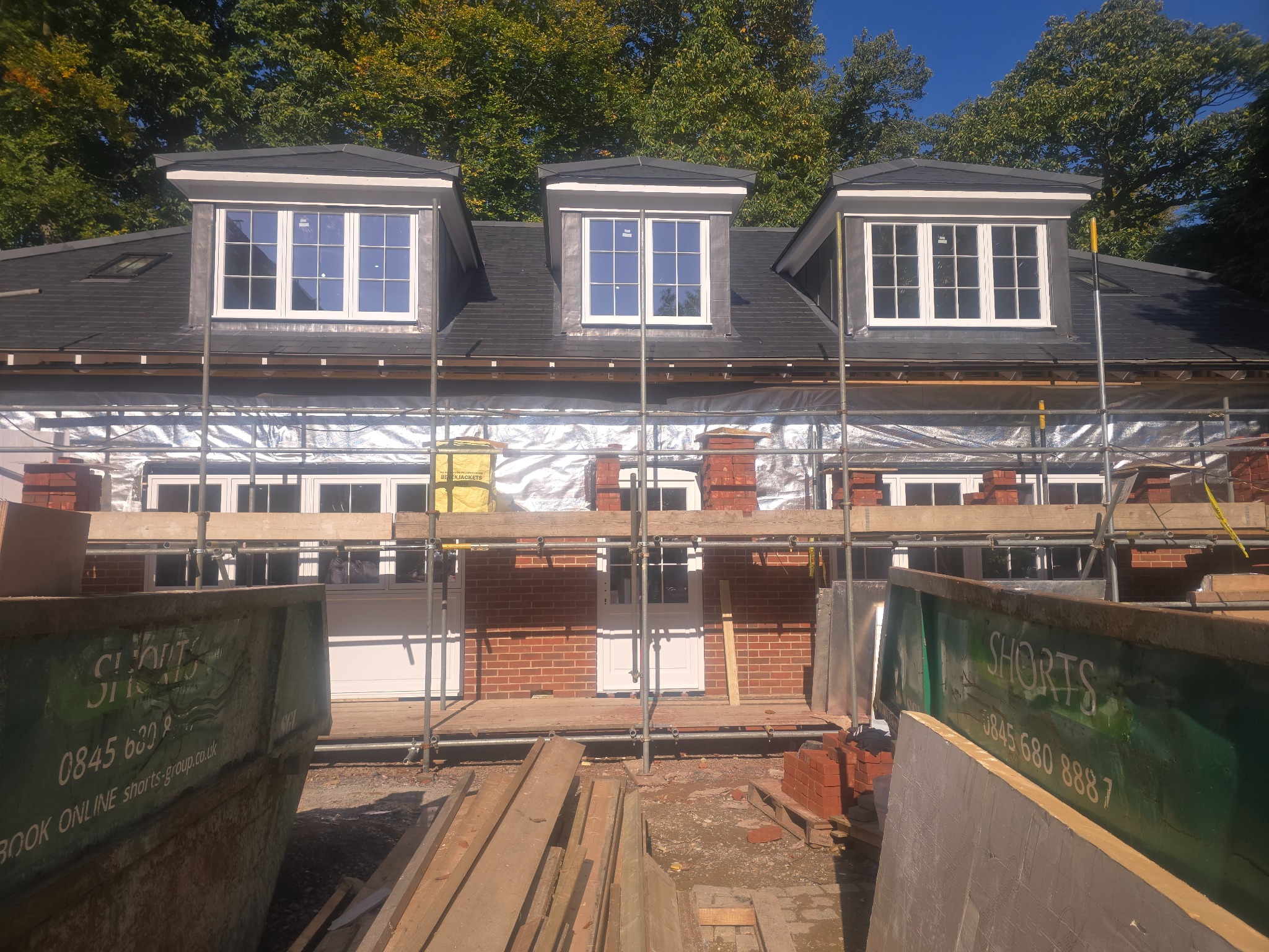 Dom w trakcie budowy z trzema oknami dachowymi i rusztowaniem. Widoczne kontenery na gruz i materiały budowlane. Częściowo ocieplona ściana budynku.