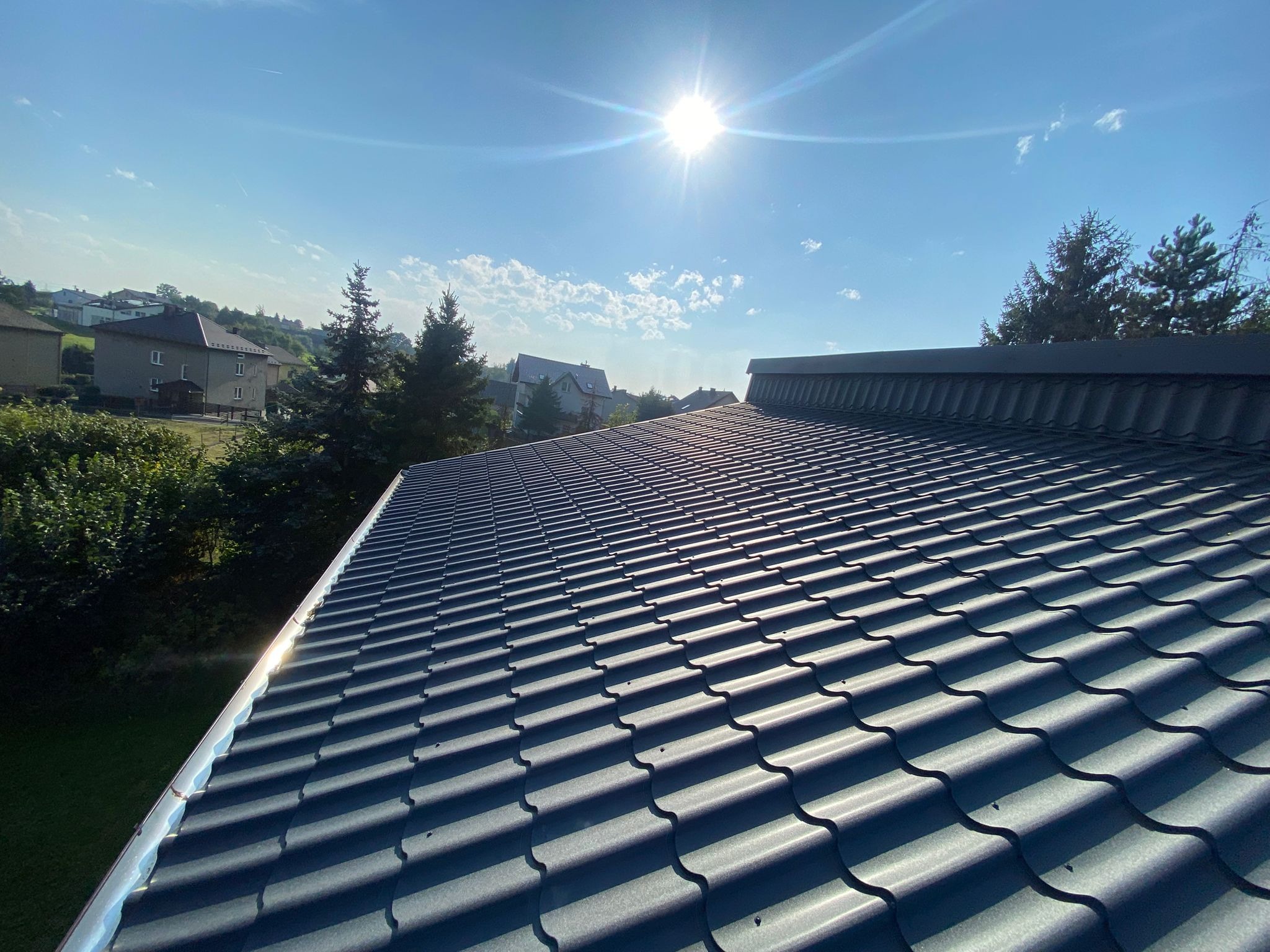 Nowy, grafitowy dach z blachodachówki w promieniach słońca, z widoczną rynną i fragmentem zabudowy w tle. Ujęcie z dołu, podkreślające regularność wzoru.