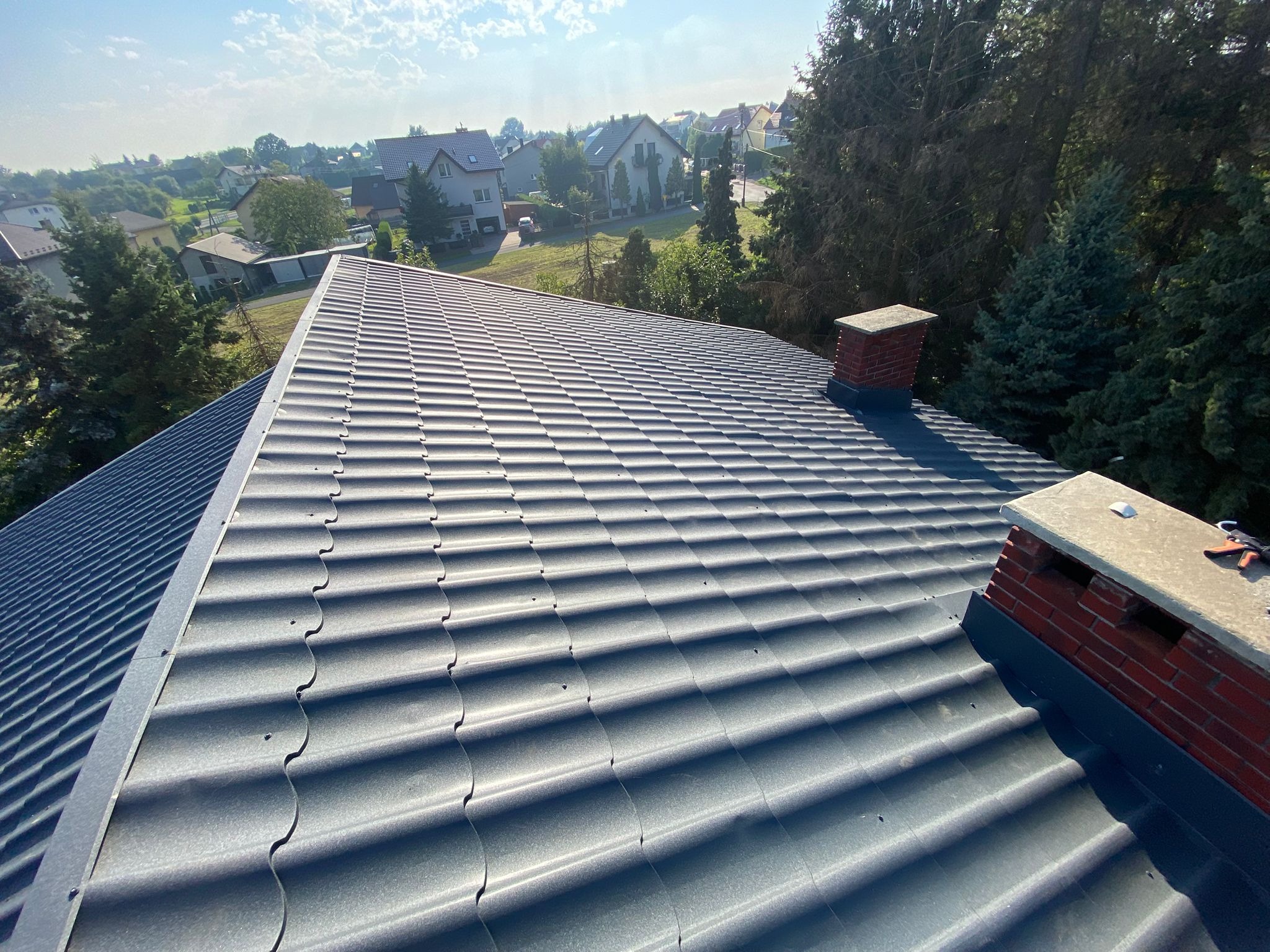 Szary dach z blachodachówki w świetle słonecznym, widok z góry na tle zabudowy jednorodzinnej i zieleni. Widoczne kominy i narzędzia dekarskie.