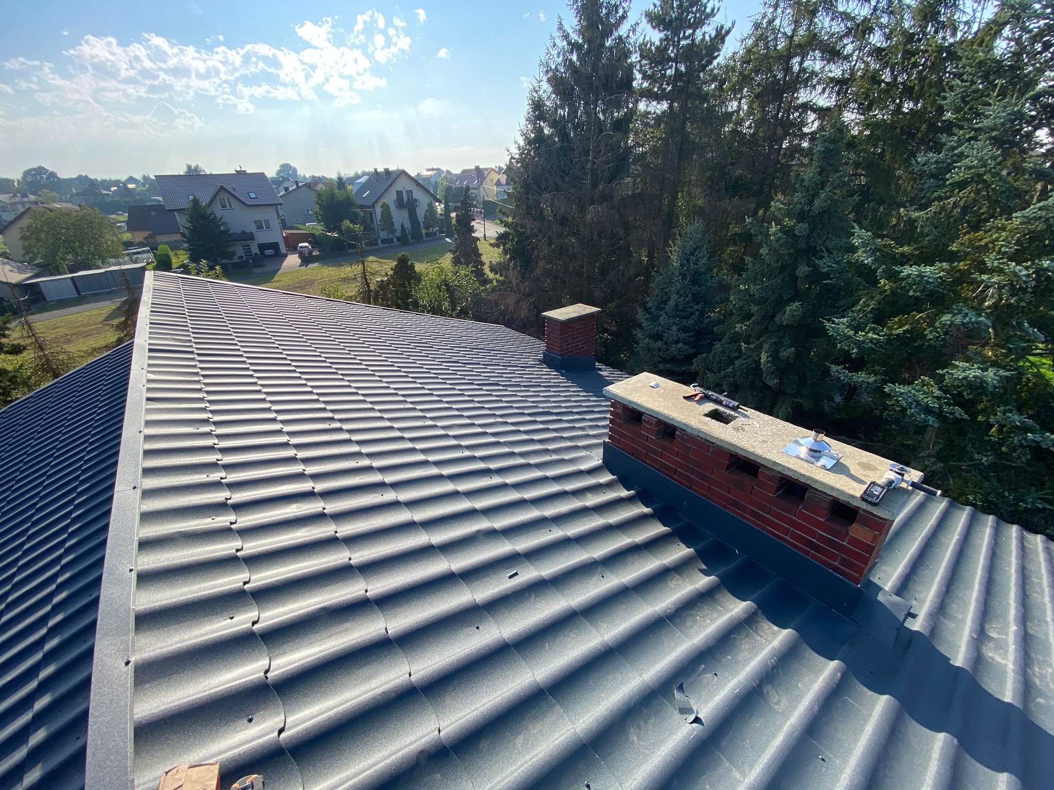 Szary dach z blachodachówki trapezowej, widok z góry na tle zieleni i zabudowań. Kominy z cegły z elementami montażowymi. Słoneczny dzień.