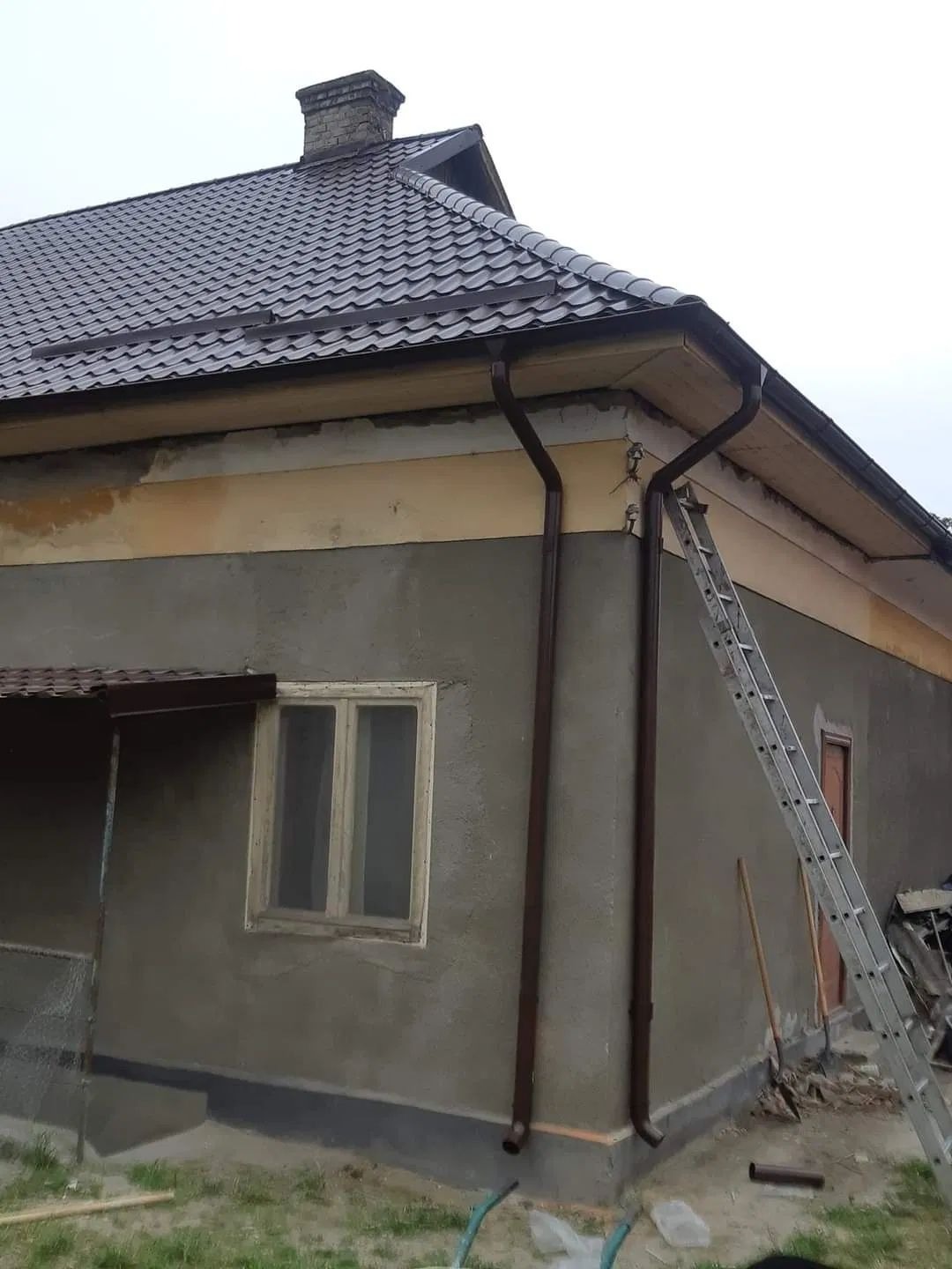 Brązowe rynny zamontowane na szarym domu z brązowym dachem. Aluminiowa drabina oparta o ścianę. Widok na narożnik budynku w pochmurny dzień.