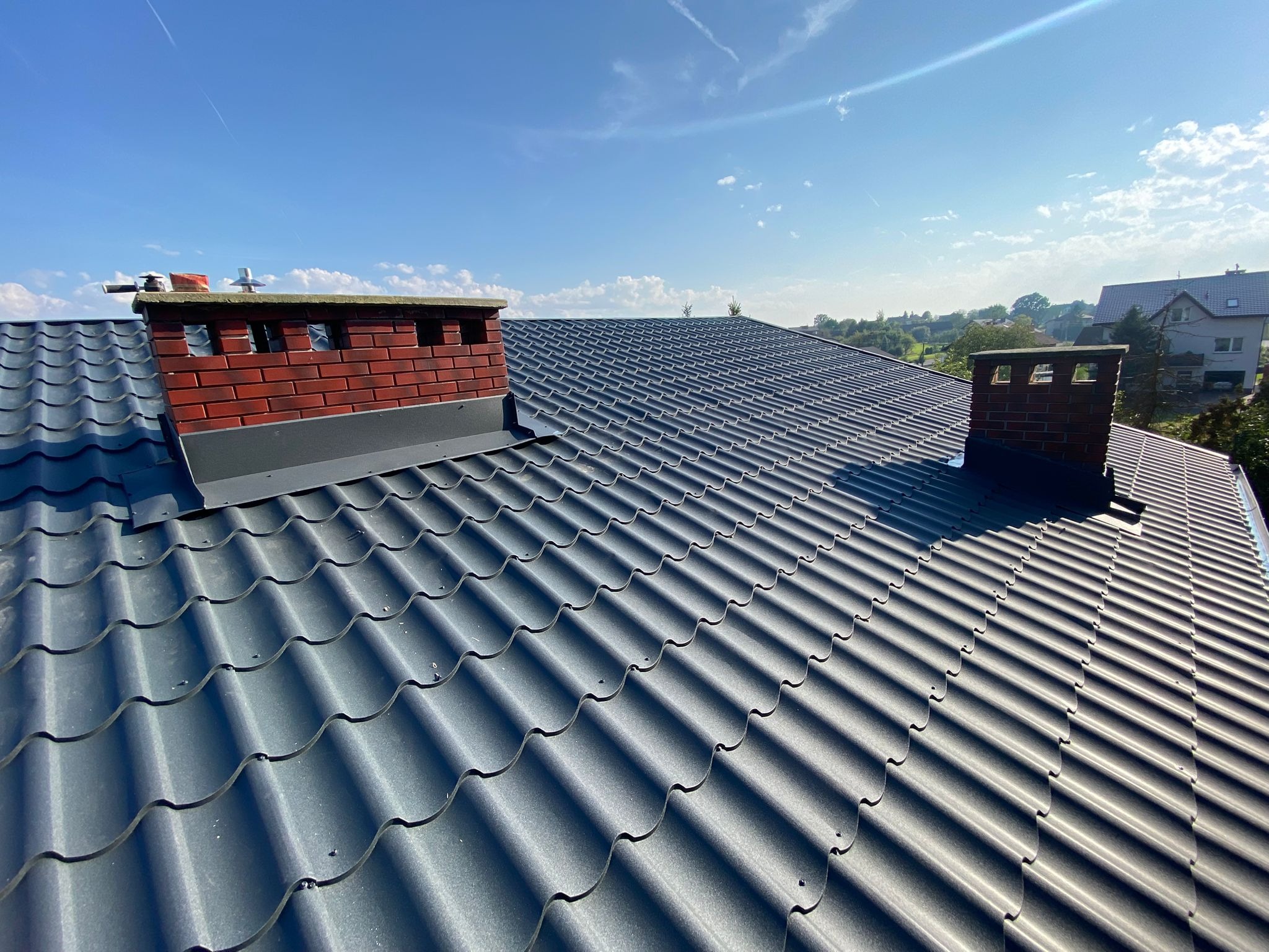 Szary dach z blachy trapezowej, dwa kominy z czerwonej cegły, błękitne niebo. Widok z góry na nowo pokryty dach domu jednorodzinnego w słoneczny dzień.