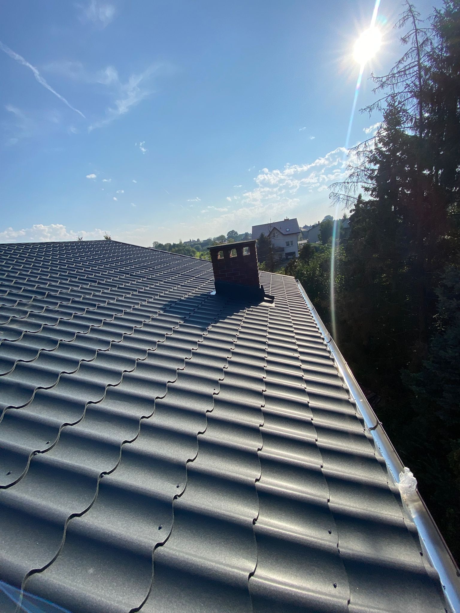 Szary dach z blachodachówki trapezowej, widok z góry, z zamontowaną rynną w słoneczny dzień. W tle drzewa i zabudowania.