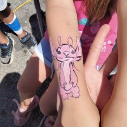 F&M EVENTS Łukasz Czendlik - Różowy, ręcznie malowany wzór Angel na przedramieniu dziecka, widoczne fragmenty ubrania i otoczenia, wykonany podczas imprezy plenerowej.