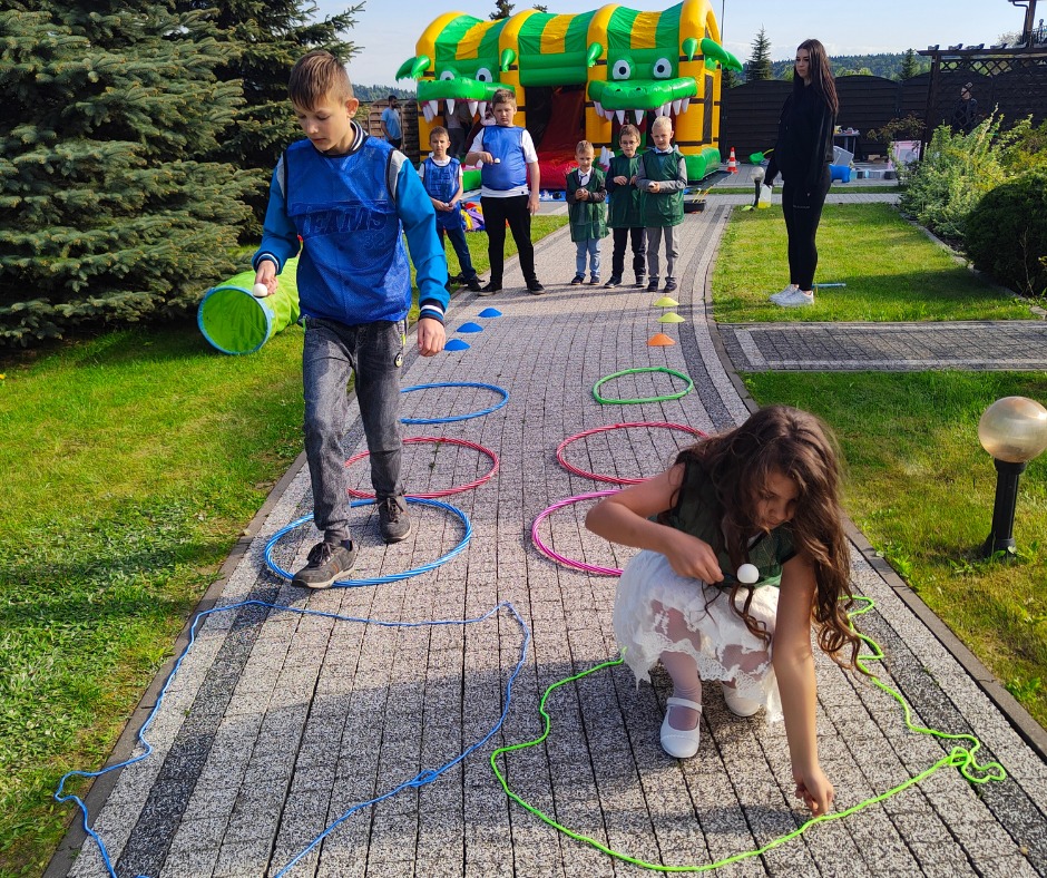 Dzieci biorą udział w grze zręcznościowej na świeżym powietrzu, z dmuchanym zamkiem w tle. Uczestnicy pokonują tor przeszkód z obręczami i tunelami.