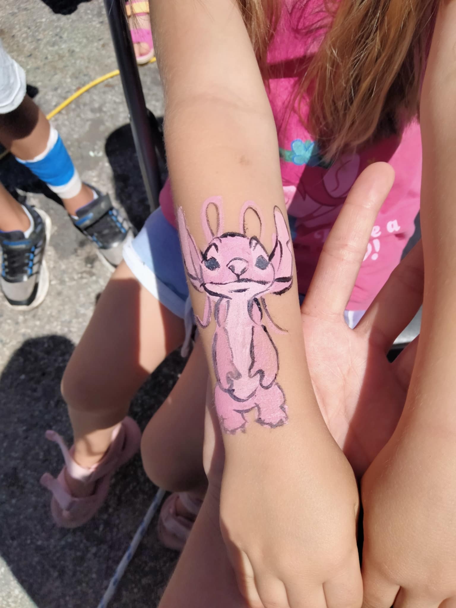 Różowy, ręcznie malowany wzór Angel na przedramieniu dziecka, widoczne fragmenty ubrania i otoczenia, wykonany podczas imprezy plenerowej.