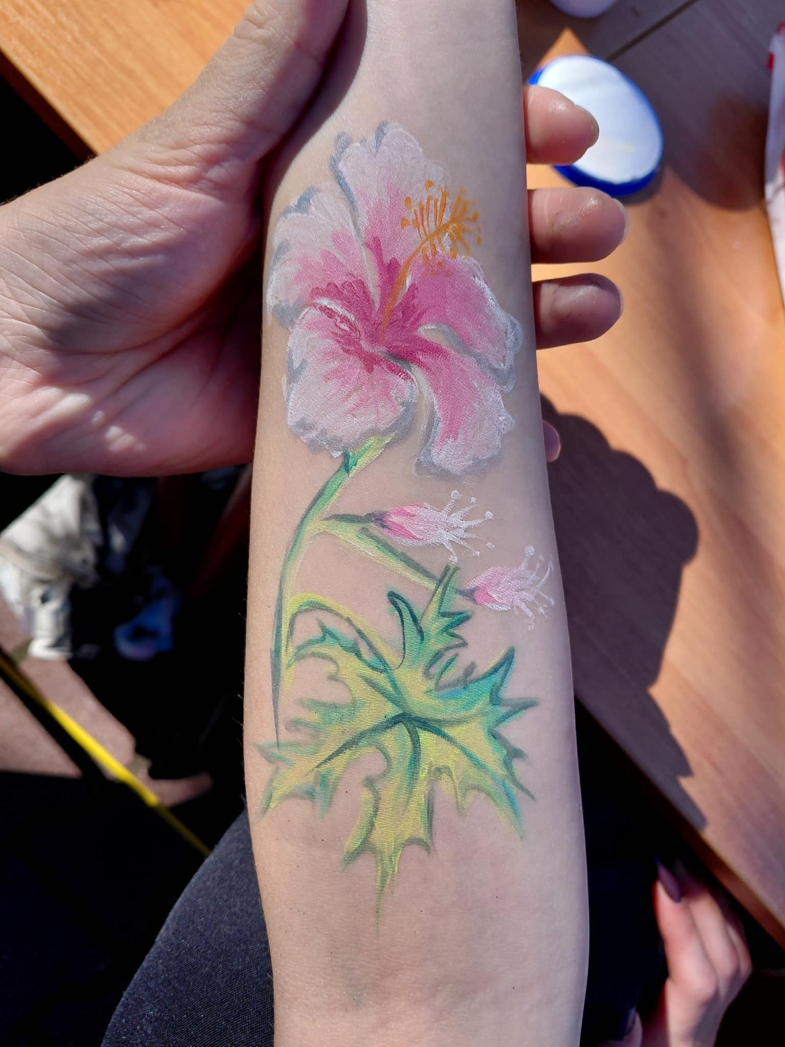 Ręka z malunkiem kwiatu hibiskusa w odcieniach różu i zieleni, wykonanym farbami do ciała. Widoczne detale pędzla i faktura skóry.