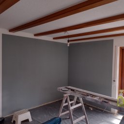 Król Pro - Pokój po malowaniu ścian na szaro, widoczne drewniane belki na suficie, drabina malarska z narzędziami i folia ochronna na podłodze.