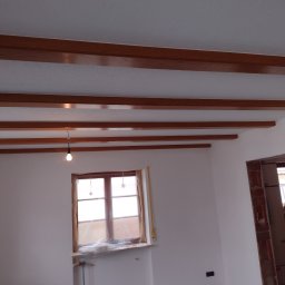 Król Pro - Białe ściany po malowaniu z drewnianymi belkami stropowymi i zabezpieczonym oknem folią malarską. Widoczny fragment instalacji grzewczej i czarna folia na podłodze.