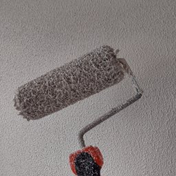 Król Pro - Wałek malarski z farbą na tle ściany o wyraźnej fakturze. Widoczne ślady świeżego malowania, narzędzie pracy w trakcie użytkowania, zbliżenie na detal faktury.