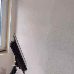 Król Pro - Ściana w trakcie wygładzania szpachelką na teleskopowym kiju. Widoczna faktura ściany przed i po nałożeniu gładzi. Okno w tle.