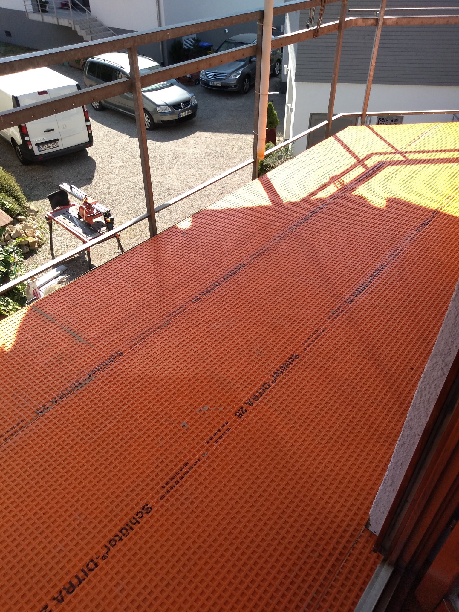 Pomarańczowa mata drenażowa Schlüter-DITRA 25 ułożona na balkonie, widok z góry. W tle samochody i narzędzia glazurnicze na rusztowaniu.