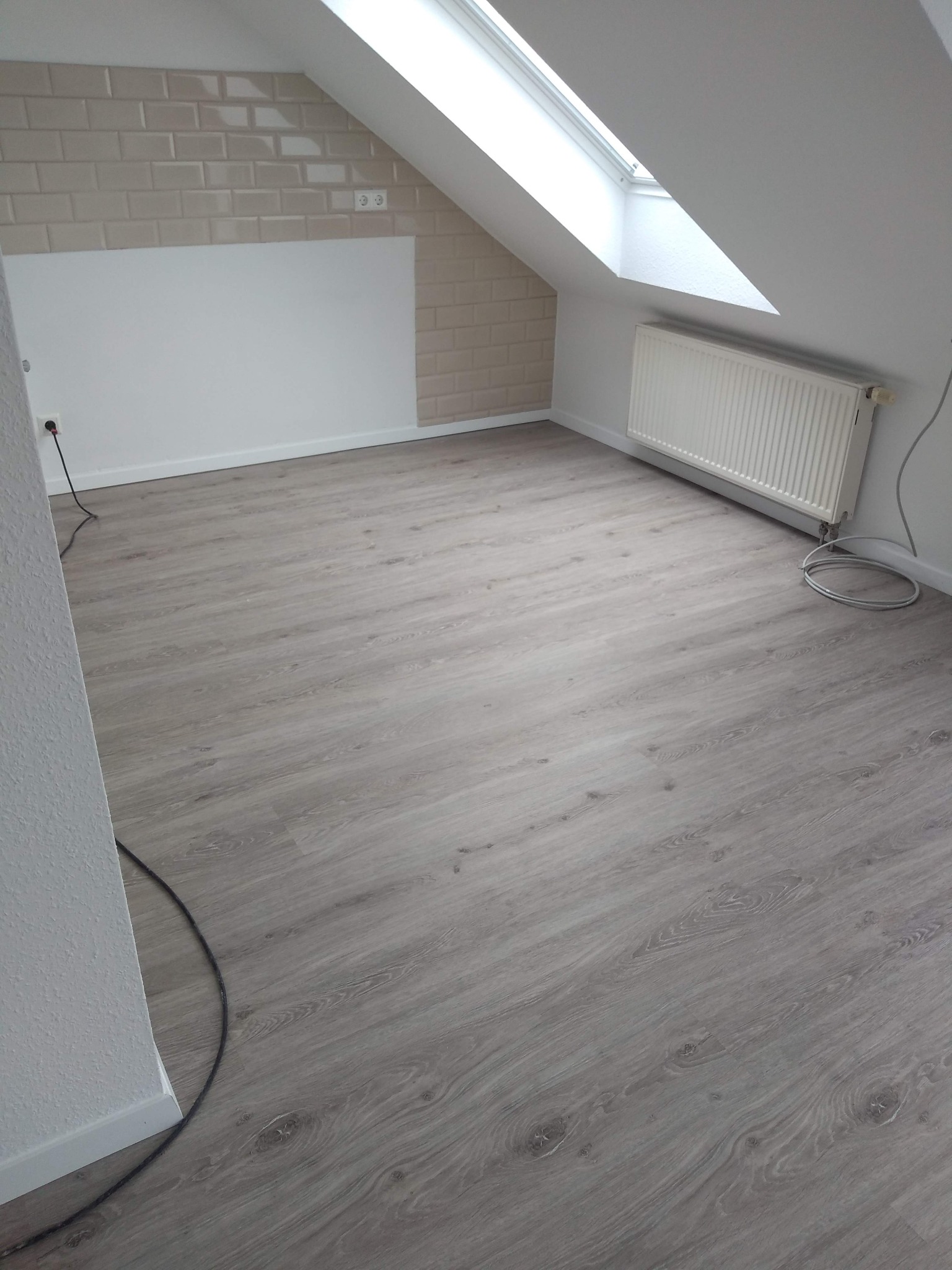 Jasne panele podłogowe w pokoju na poddaszu z oknem dachowym. Ściany częściowo wyłożone beżowymi płytkami imitującymi cegłę, grzejnik pod oknem.