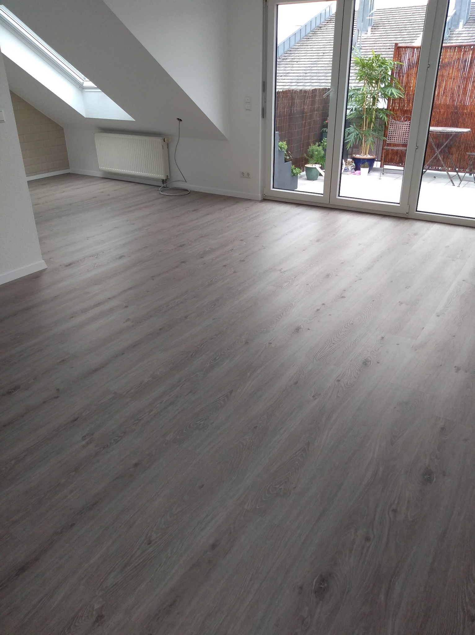 Jasne, przestronne poddasze z panelami podłogowymi w odcieniach szarości. Okno dachowe i duże okno balkonowe wpuszczają dużo światła. Widok na taras z roślinami.