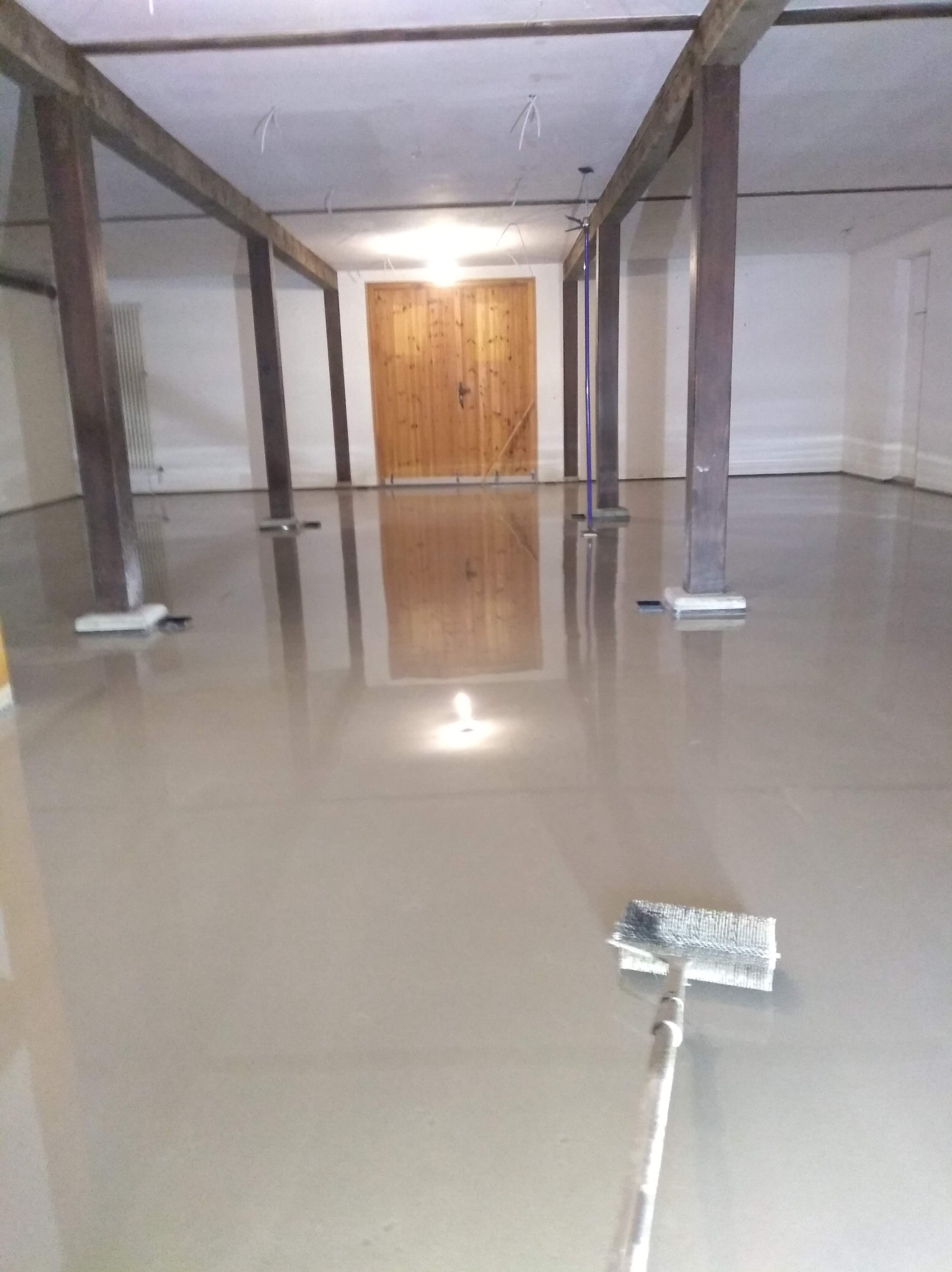 Wylewka samopoziomująca w pomieszczeniu z drewnianą konstrukcją stropu. Widoczny wałek do rozprowadzania masy. Gładka, jasna powierzchnia podłogi.