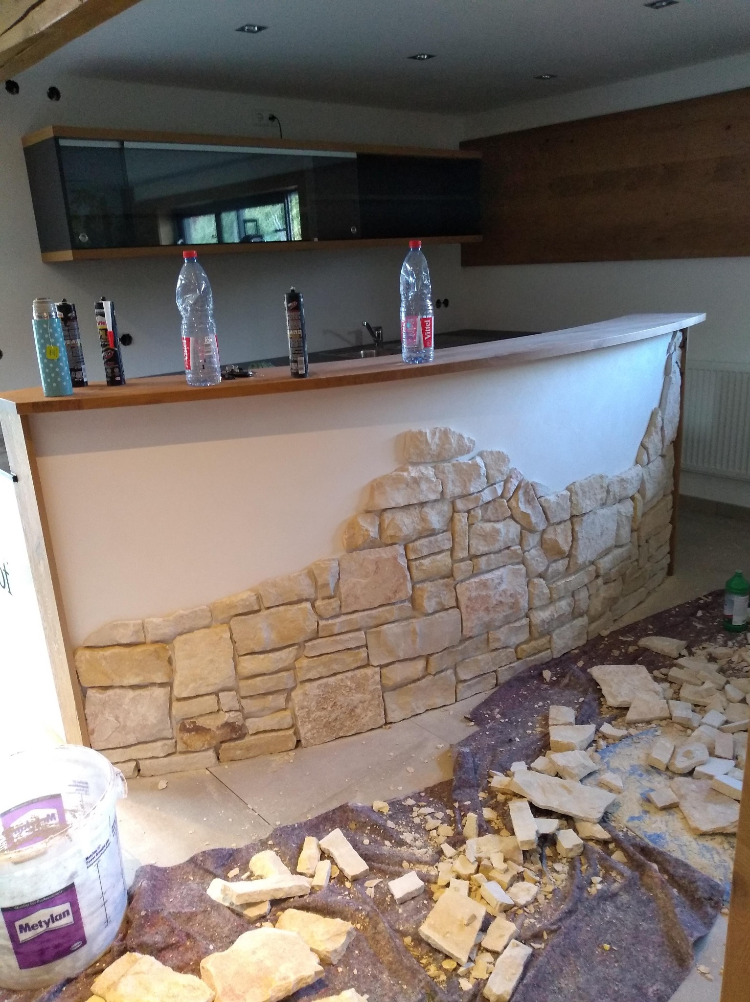 Bar z częściowo wykończoną okładziną z kamienia dekoracyjnego. Widoczne narzędzia i materiały budowlane. W tle szafki i elementy wyposażenia wnętrza.