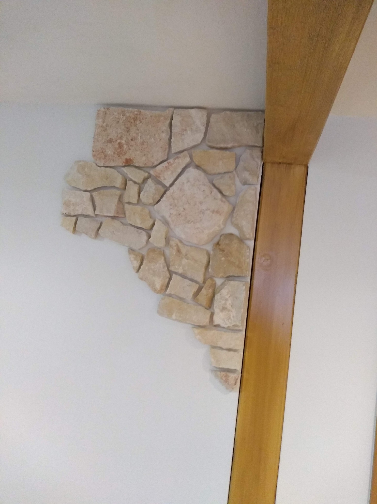 Dekoracyjna ściana z nieregularnych, jasnych kamieni, przylegająca do drewnianej belki w rogu pomieszczenia. Biała ściana, ciepłe oświetlenie.