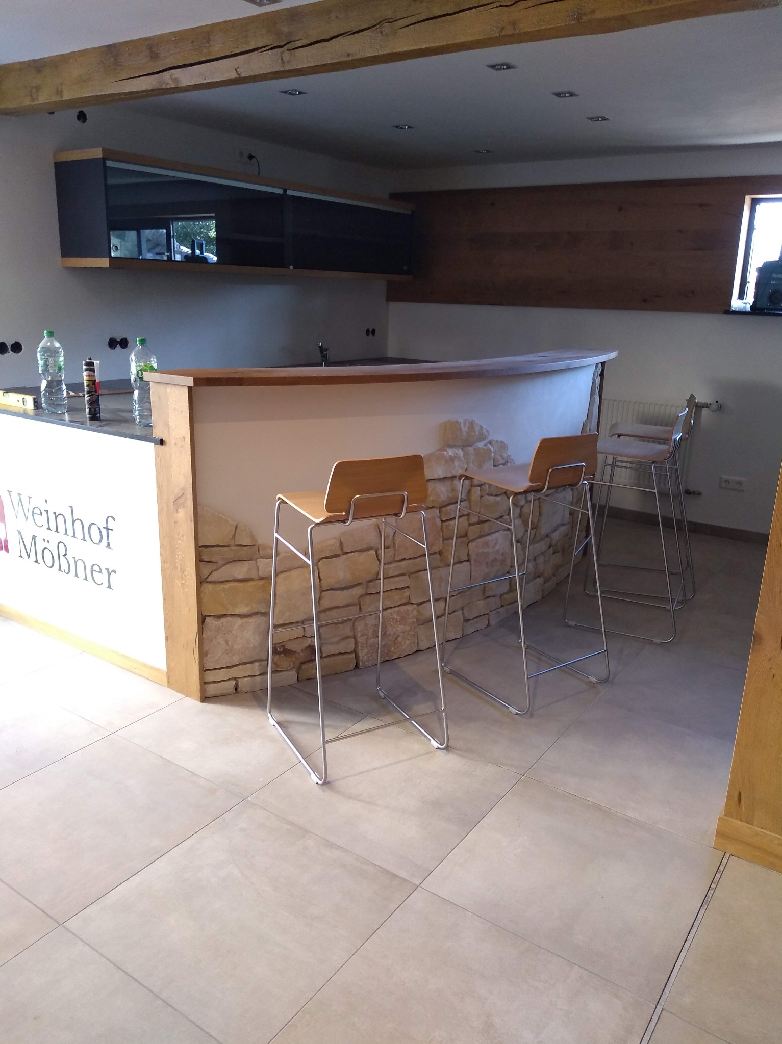 Bar z kamienną okładziną i drewnianym blatem, trzy krzesła barowe, płytki podłogowe w odcieniach beżu. Widoczne elementy drewniane na suficie i ścianie.