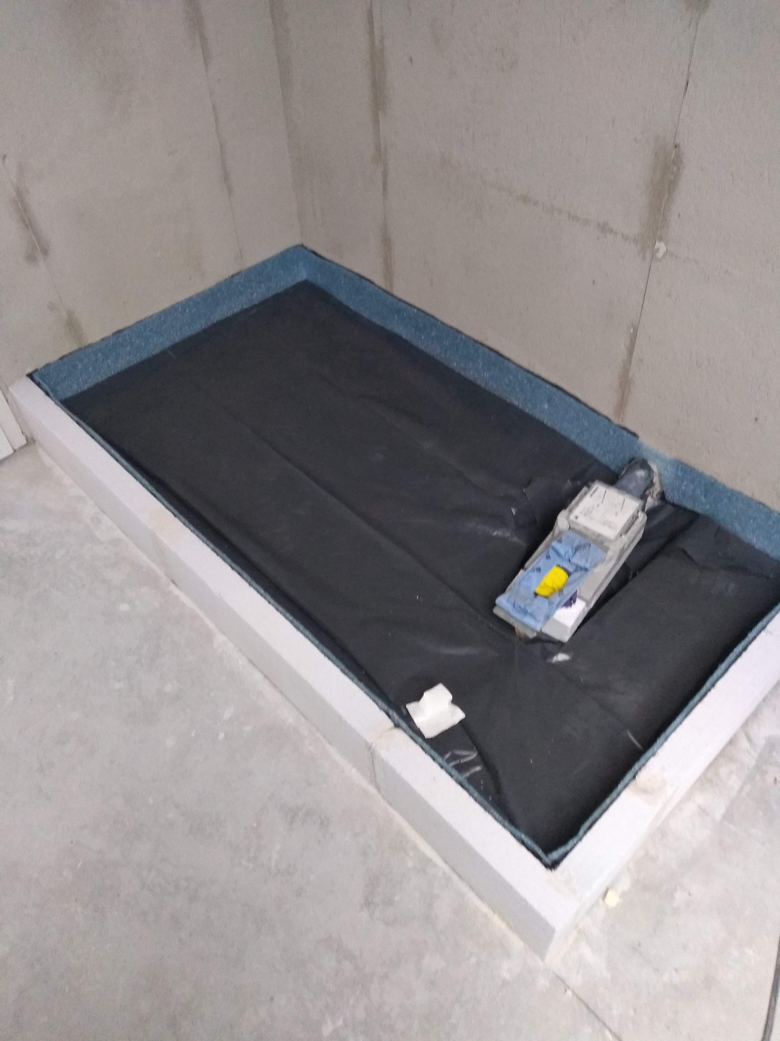 Przygotowany brodzik z izolacją i folią w narożniku łazienki przed dalszymi pracami wykończeniowymi. Widoczny poziomica i fragment ściany w surowym stanie.