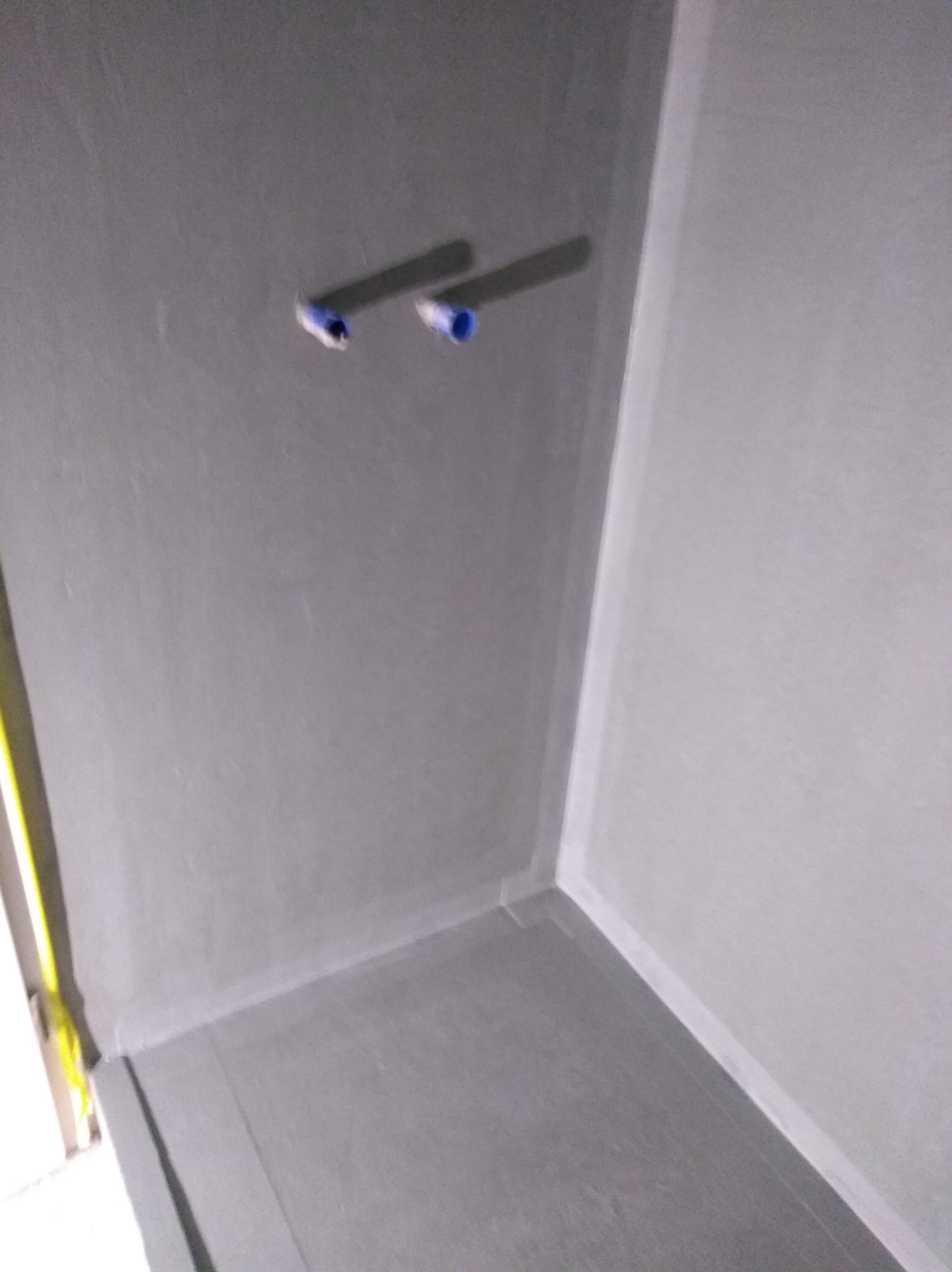 Szara, świeżo wykończona łazienka z widocznymi rurami instalacji wodnej wystającymi ze ściany. Minimalistyczne wykończenie, surowe ściany i podłoga.