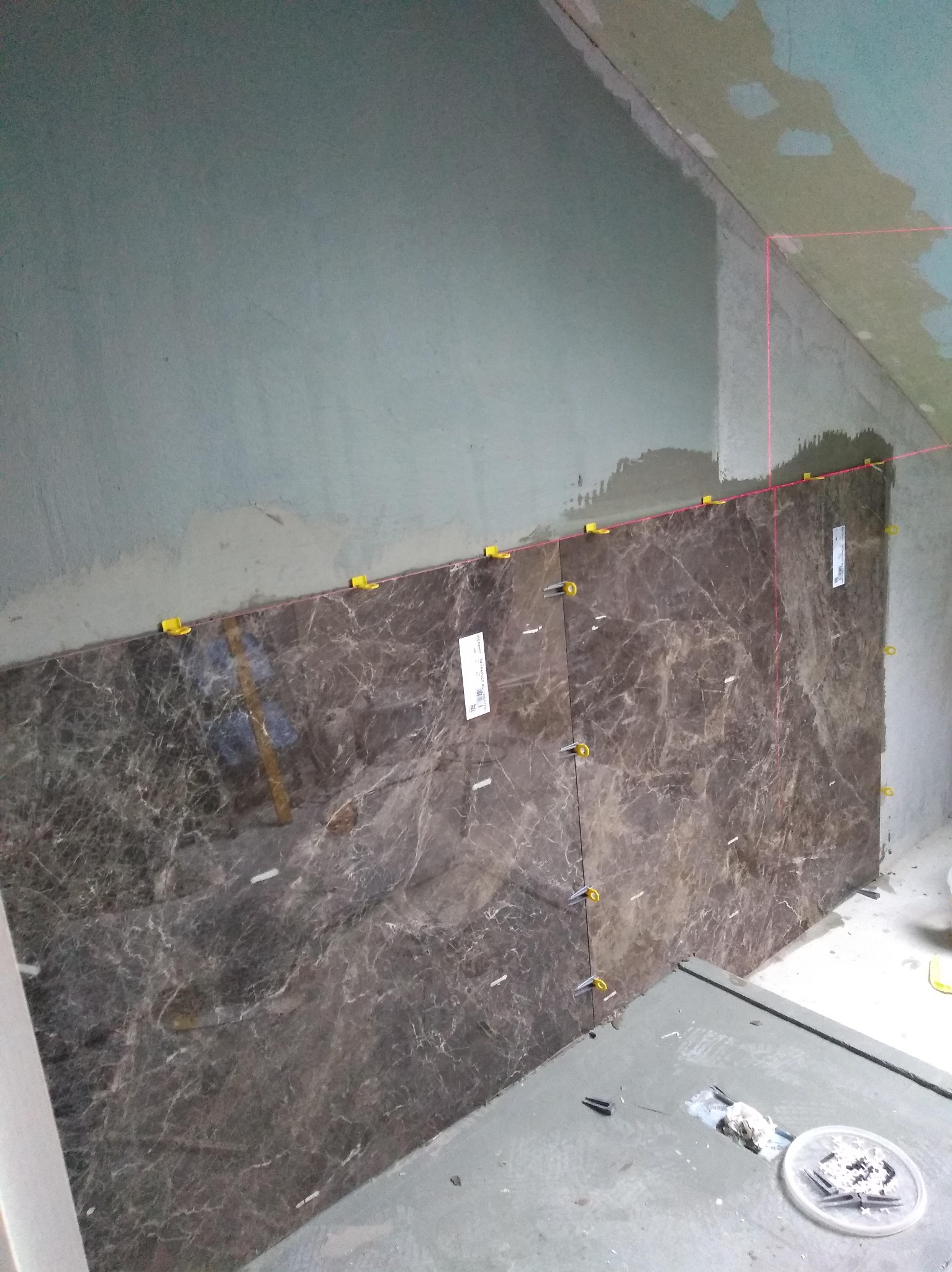 Układanie płytek imitujących marmur na ścianie łazienki, z użyciem poziomicy laserowej i klipsów poziomujących. Widoczne narzędzia glazurnicze.