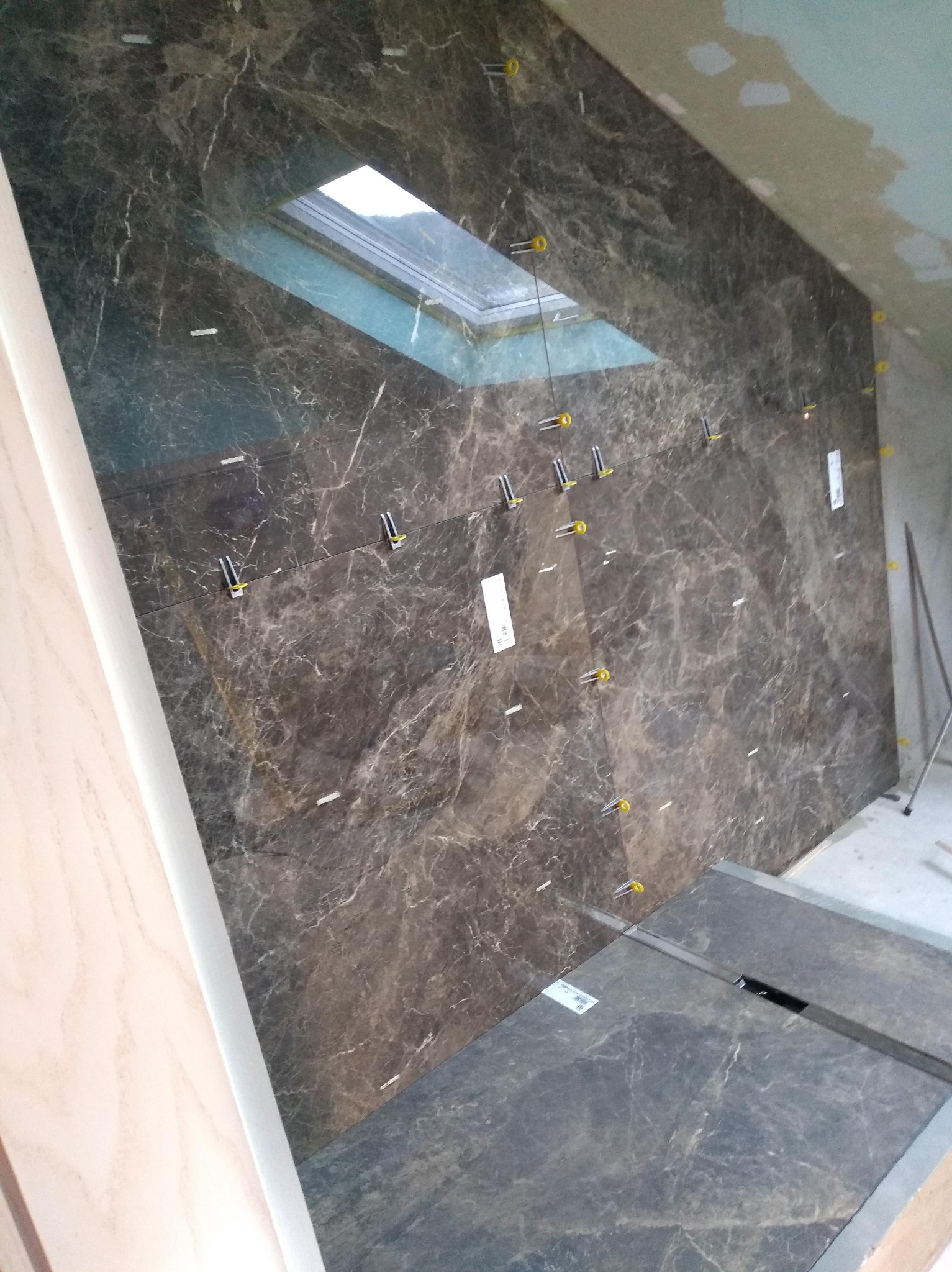 Układanie płytek imitujących marmur na ścianie łazienki ze świetlikiem dachowym. Widoczne narzędzia glazurnicze i poziomica. Prace w toku.