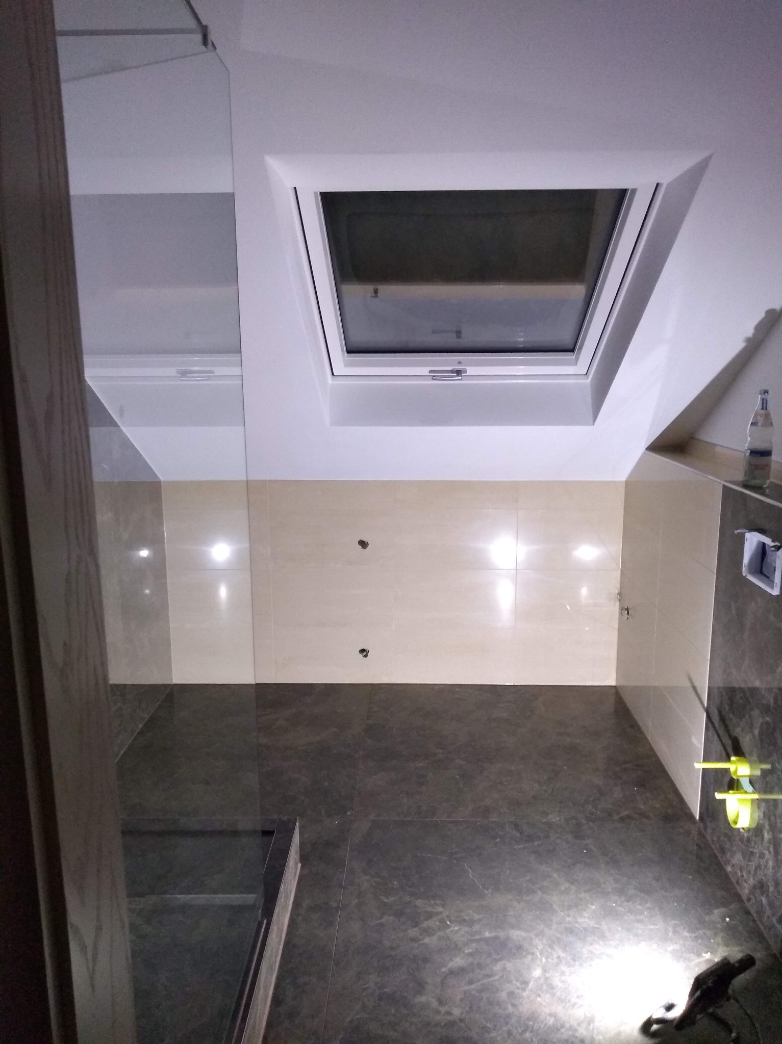 Wykończona łazienka z oknem dachowym, beżowe i grafitowe płytki, szklana kabina prysznicowa. Widok z góry, oświetlenie punktowe.