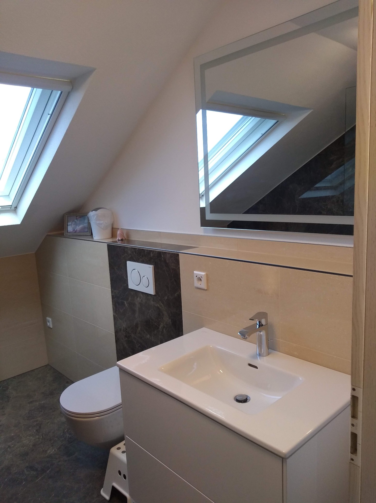 Jasna łazienka na poddaszu z oknem dachowym, beżowe płytki, umywalka z szafką i lustro z podświetleniem. Uwagę przykuwa marmurowy akcent na ścianie.