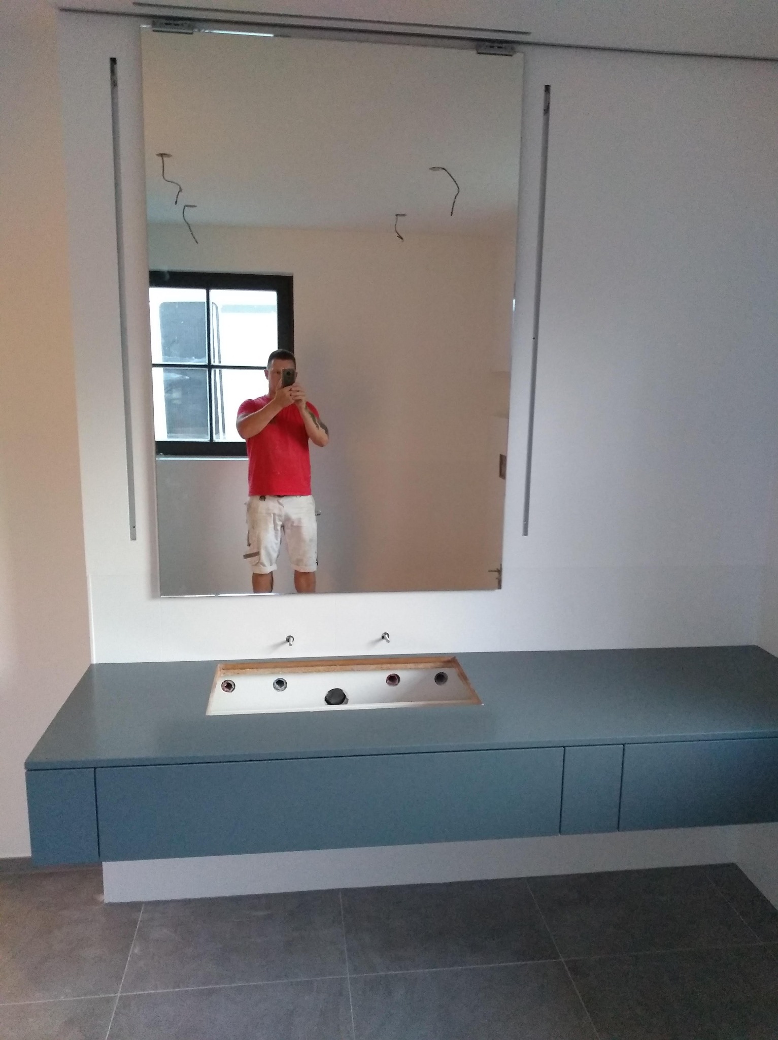 Niebieska szafka pod umywalkę z lustrem w nowoczesnej łazience. Widoczne przygotowane otwory na baterie i odpływ. W lustrze odbija się osoba robiąca zdjęcie.