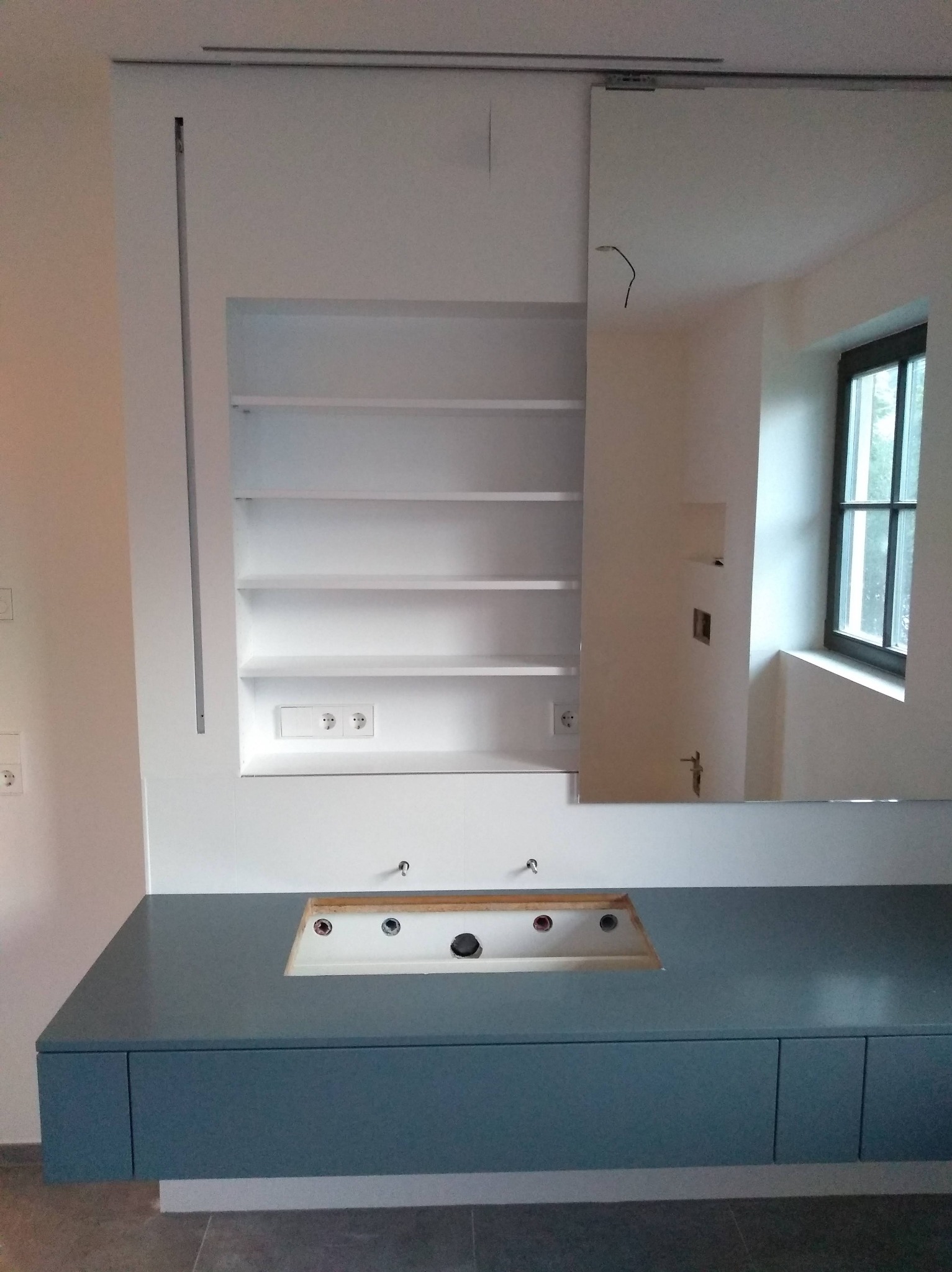 Nowoczesna łazienka w trakcie wykańczania: szafka pod umywalkę w odcieniach szarości, białe półki w zabudowie i przesuwne lustro. Widoczne gniazdka i okno.