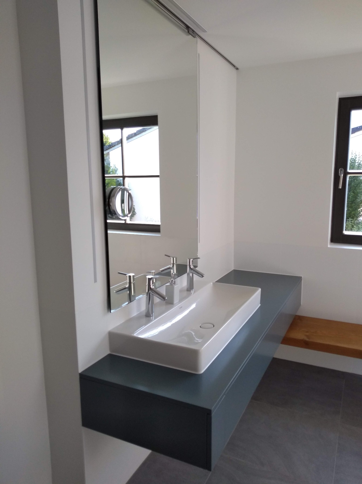 Nowoczesna łazienka: biała umywalka nablatowa z dwoma kranami na szarym blacie, duże lustro przesuwne, szare płytki podłogowe i okno z widokiem na zieleń.