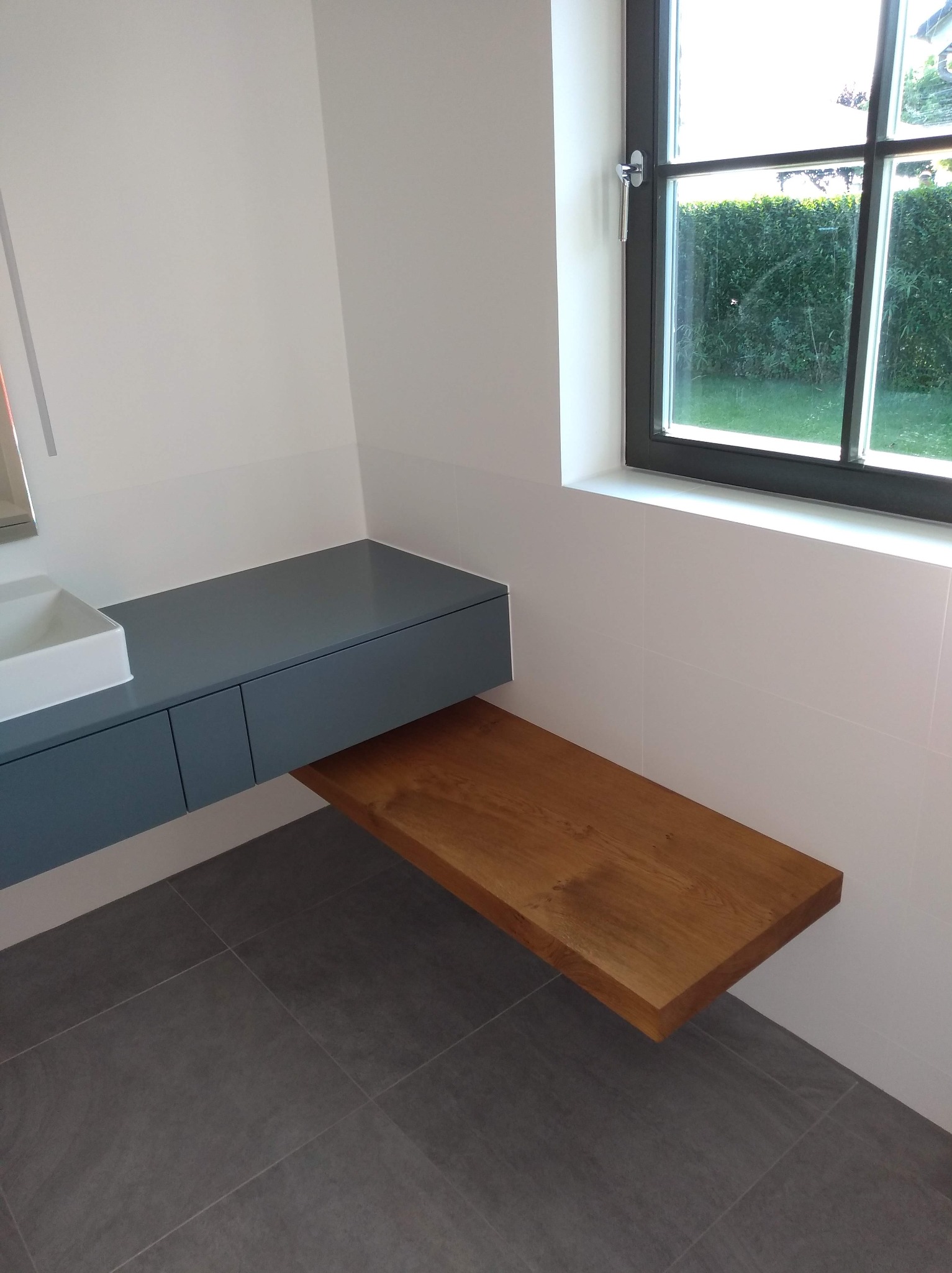 Nowoczesna łazienka z szarą szafką pod umywalką i drewnianym blatem, szare płytki podłogowe, okno z widokiem na zieleń. Minimalistyczny design, jasne wnętrze.