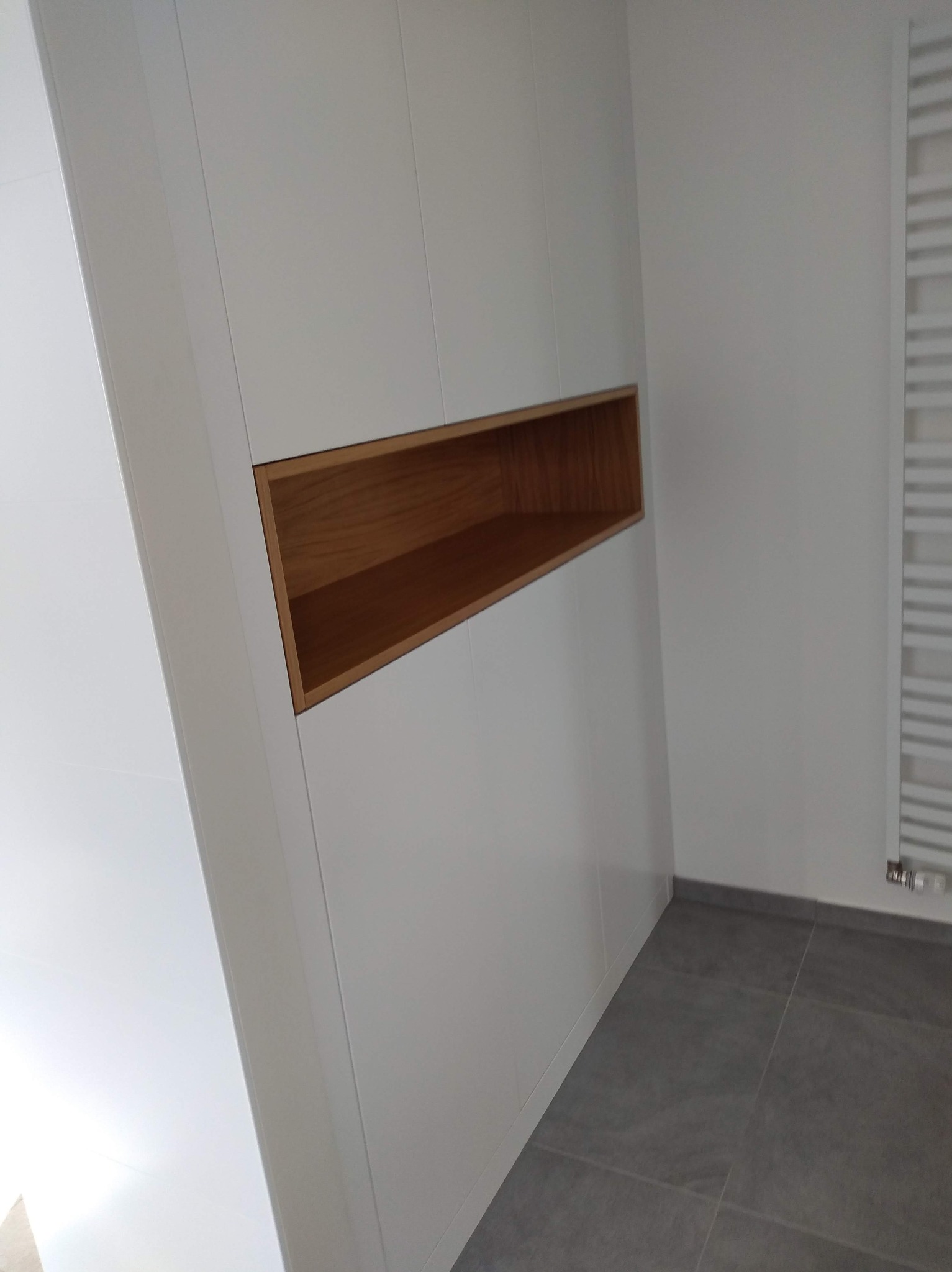 Biała zabudowa ściany z wnęką z drewna w łazience. Szare płytki podłogowe i grzejnik drabinkowy w tle. Minimalistyczny design.