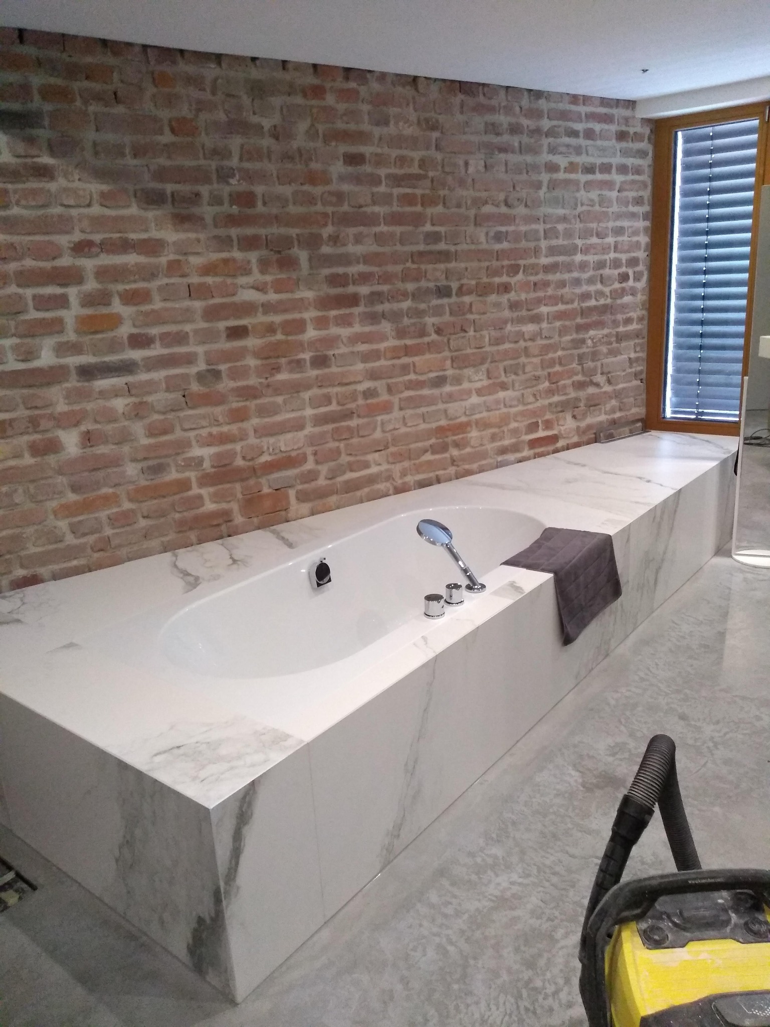 Wykończona wanna z marmurową obudową i baterią, na tle ceglanej ściany w łazience w Krotoszynie. Widoczny fragment okna z żaluzjami i sprzęt budowlany.