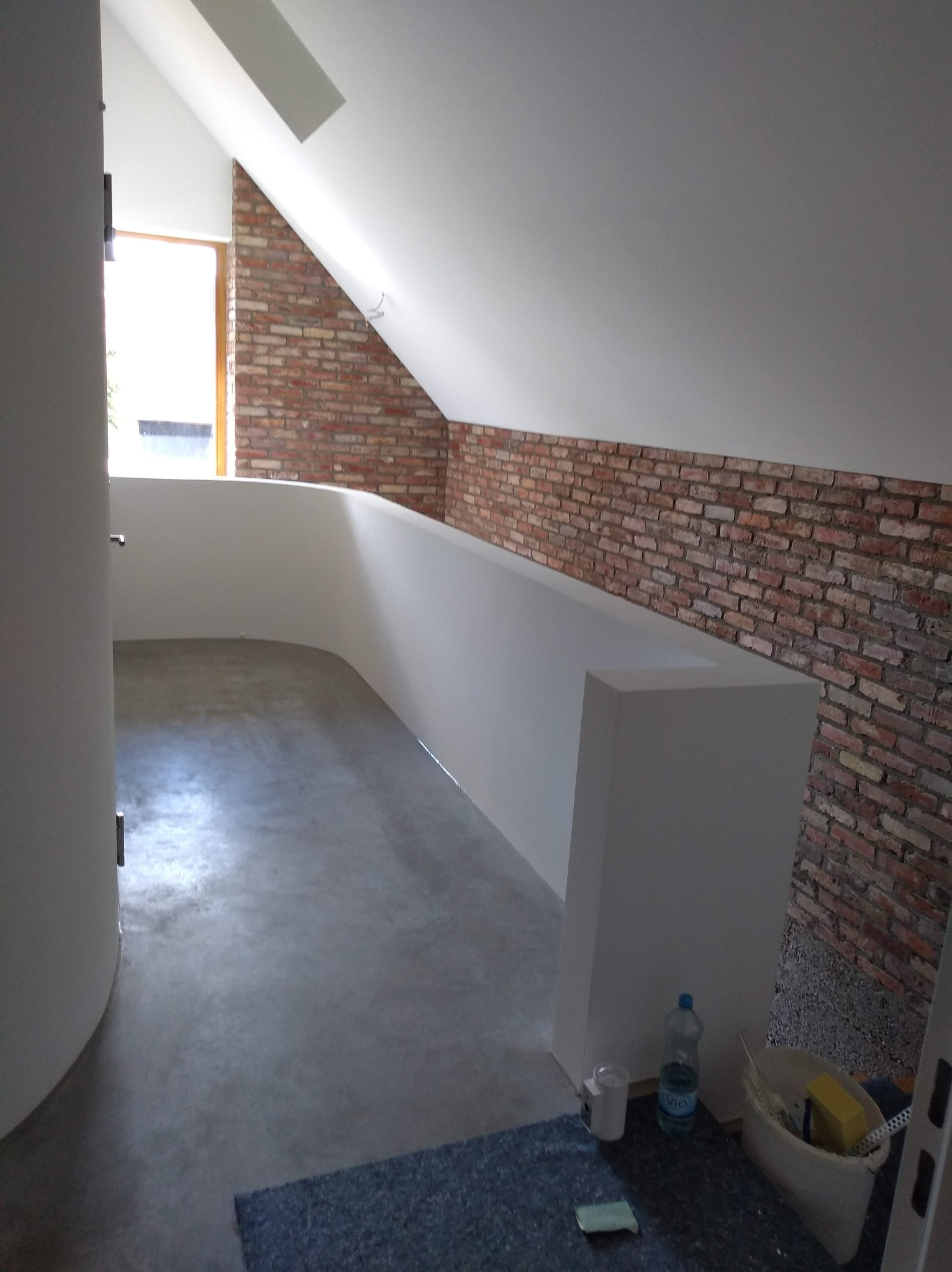 Poddasze po remoncie: białe ściany, ceglana ściana akcentowa, szara podłoga. Widoczne narzędzia malarskie i butelka wody. Ukończone wykończenie wnętrza.