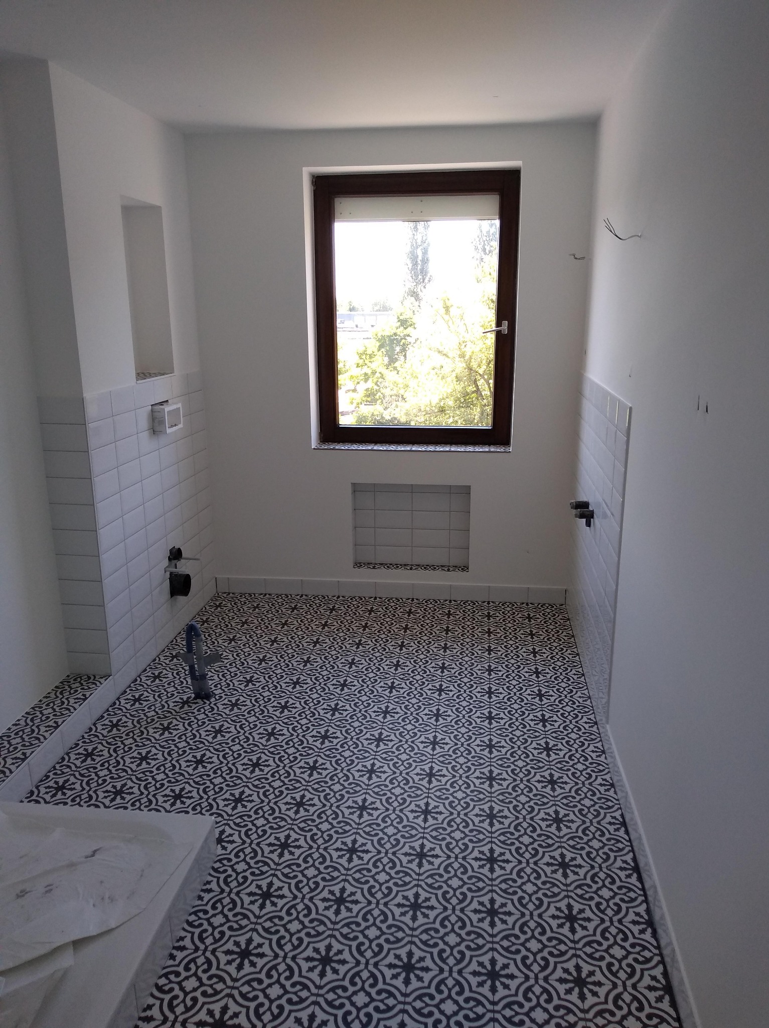 Biała łazienka z wzorzystą, czarno-białą podłogą. Ściany wyłożone białymi płytkami do połowy wysokości, okno z brązową ramą. Widoczne instalacje wodne.