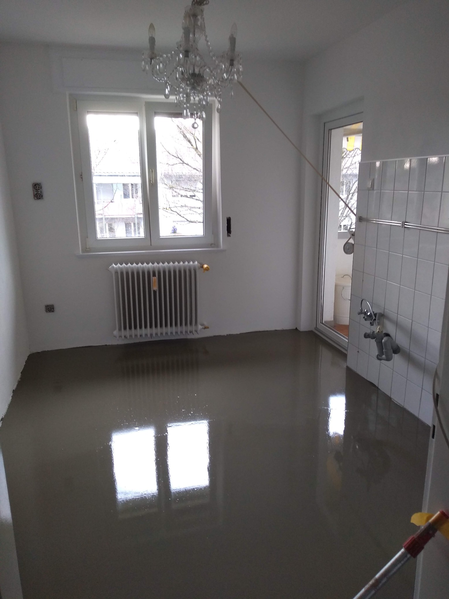 Wnętrze po wylaniu samopoziomującej wylewki. Widoczne okno z grzejnikiem, białe ściany, fragment glazury i żyrandol. Podłoga mokra, odbija światło.