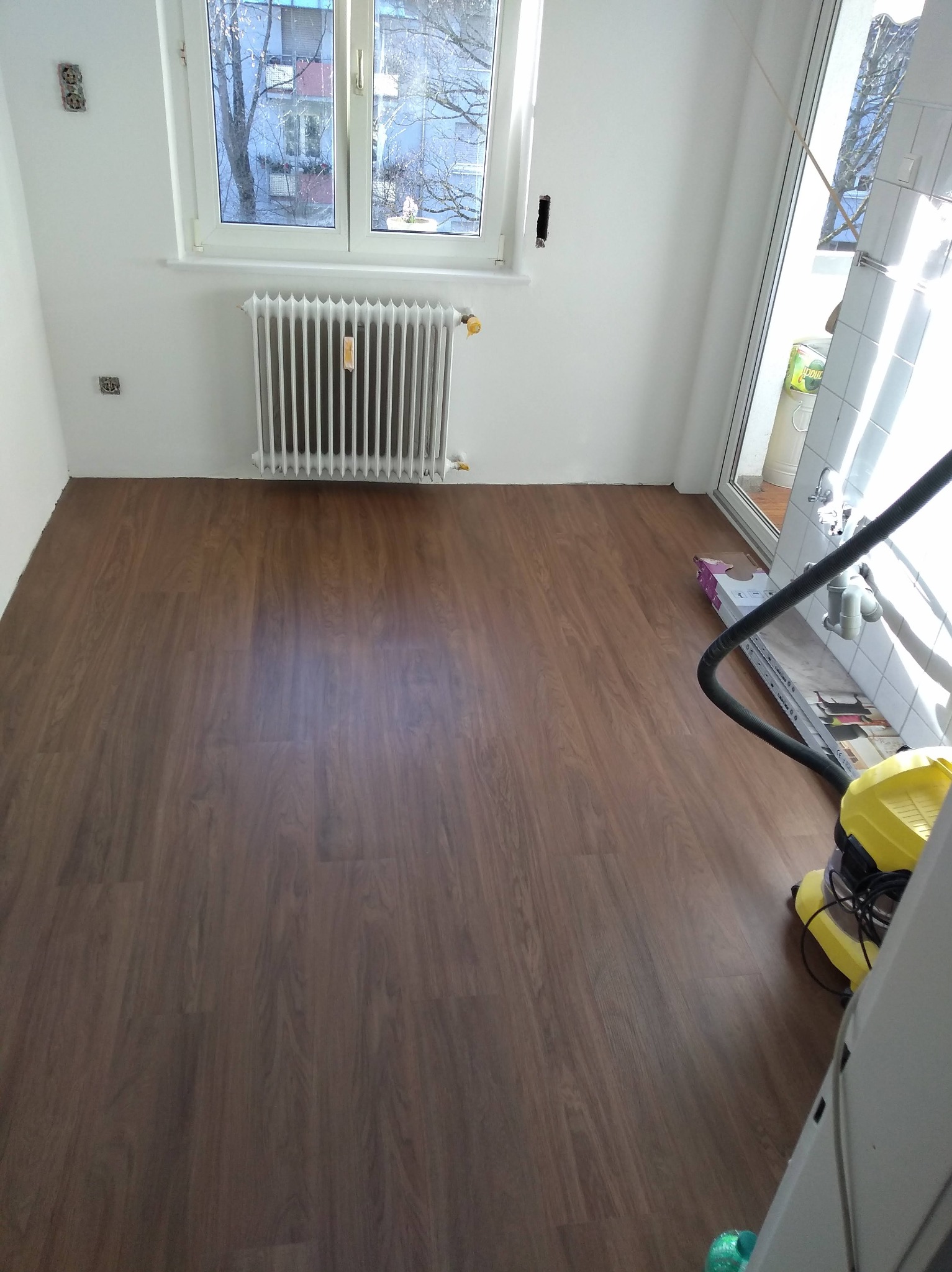 Ukończona podłoga z paneli w jasnym pokoju z grzejnikiem i oknem. Widoczny odkurzacz i resztki materiałów po montażu. Nowe panele podłogowe w odcieniach brązu.