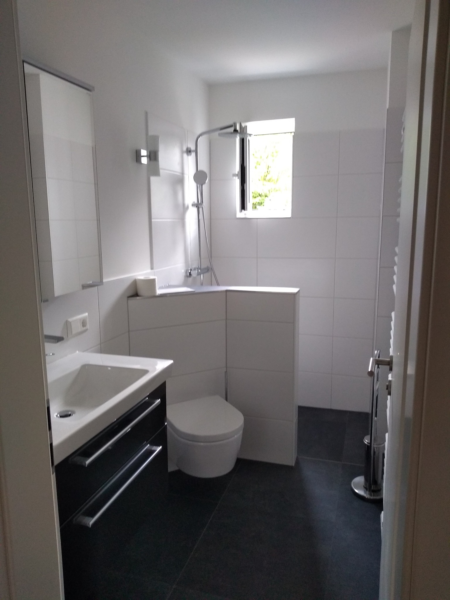 Nowoczesna łazienka z białymi płytkami, czarną podłogą i szafką pod umywalką. Widoczny fragment okna i kabina prysznicowa. Minimalistyczny design.