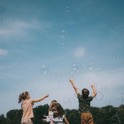 Kreatywne Urodziny Adriana Foryś - Dzieci bawią się bańkami mydlanymi na tle błękitnego nieba. Dziewczynka z boku puszcza bańki za pomocą sznurka i patyków. Radosna atmosfera zabawy na świeżym powietrzu.